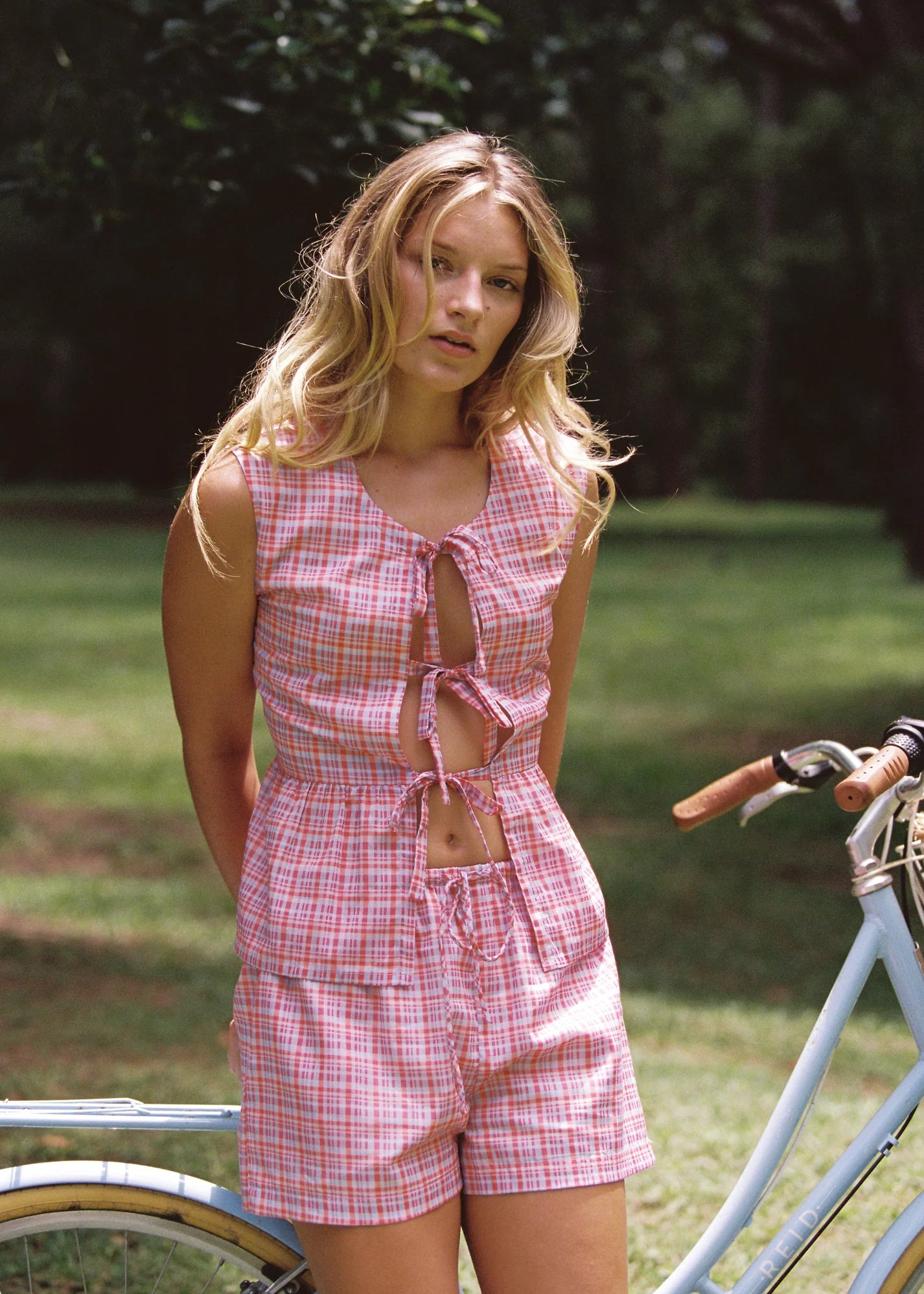 A person with long blonde hair stands in a grassy area, wearing the ethically made JUNI ~ Dylan Short, a pink plaid sleeveless romper with front ties. A vintage-style blue bicycle nearby perfectly complements the lush green foliage.