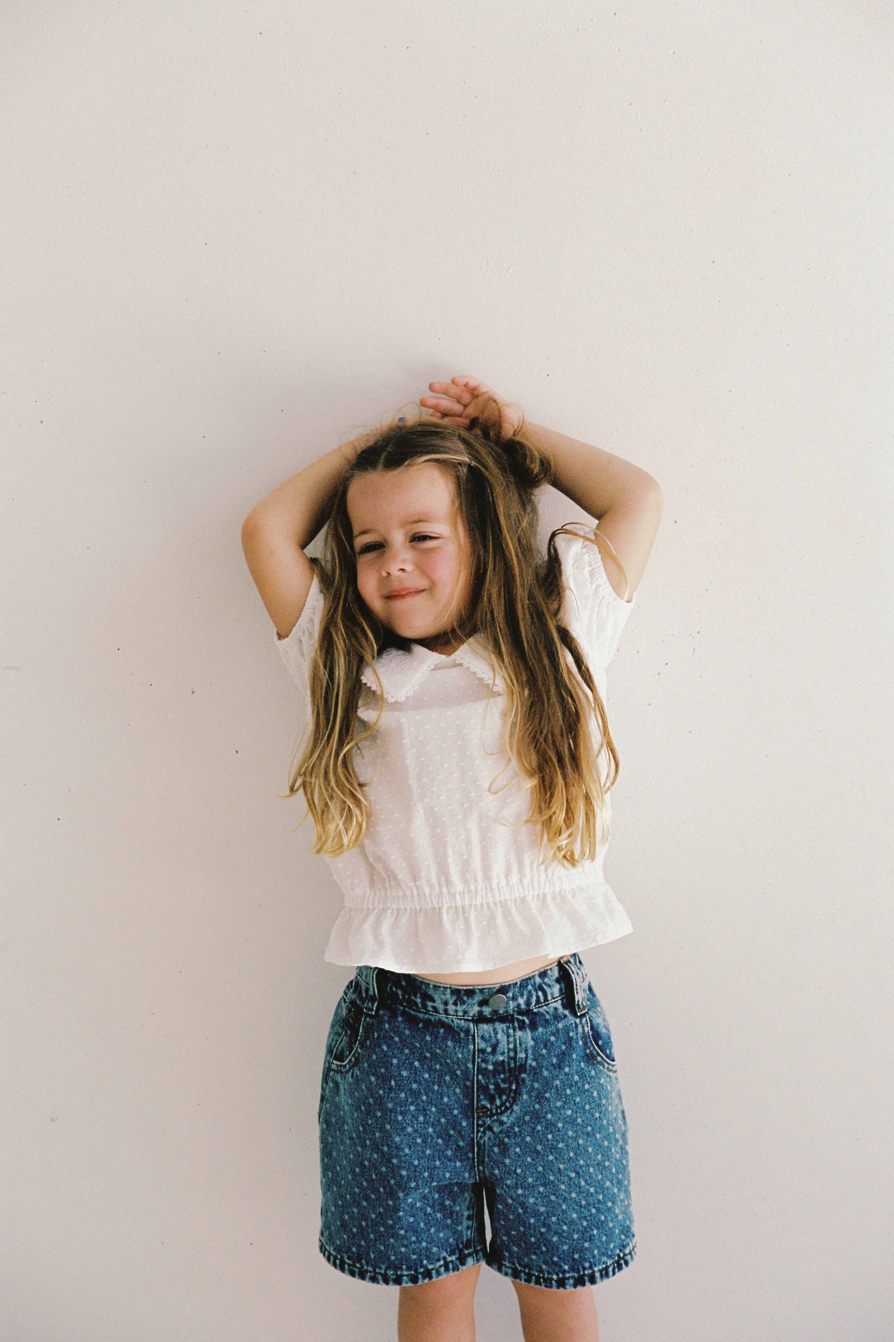 A young girl with long brown hair stands against a white wall, smiling with her arms raised above her head. She is wearing a white short-sleeved top and JUNI JUNIOR Junior Bud Denim Short Spot — blue denim shorts with an elastic waistband.