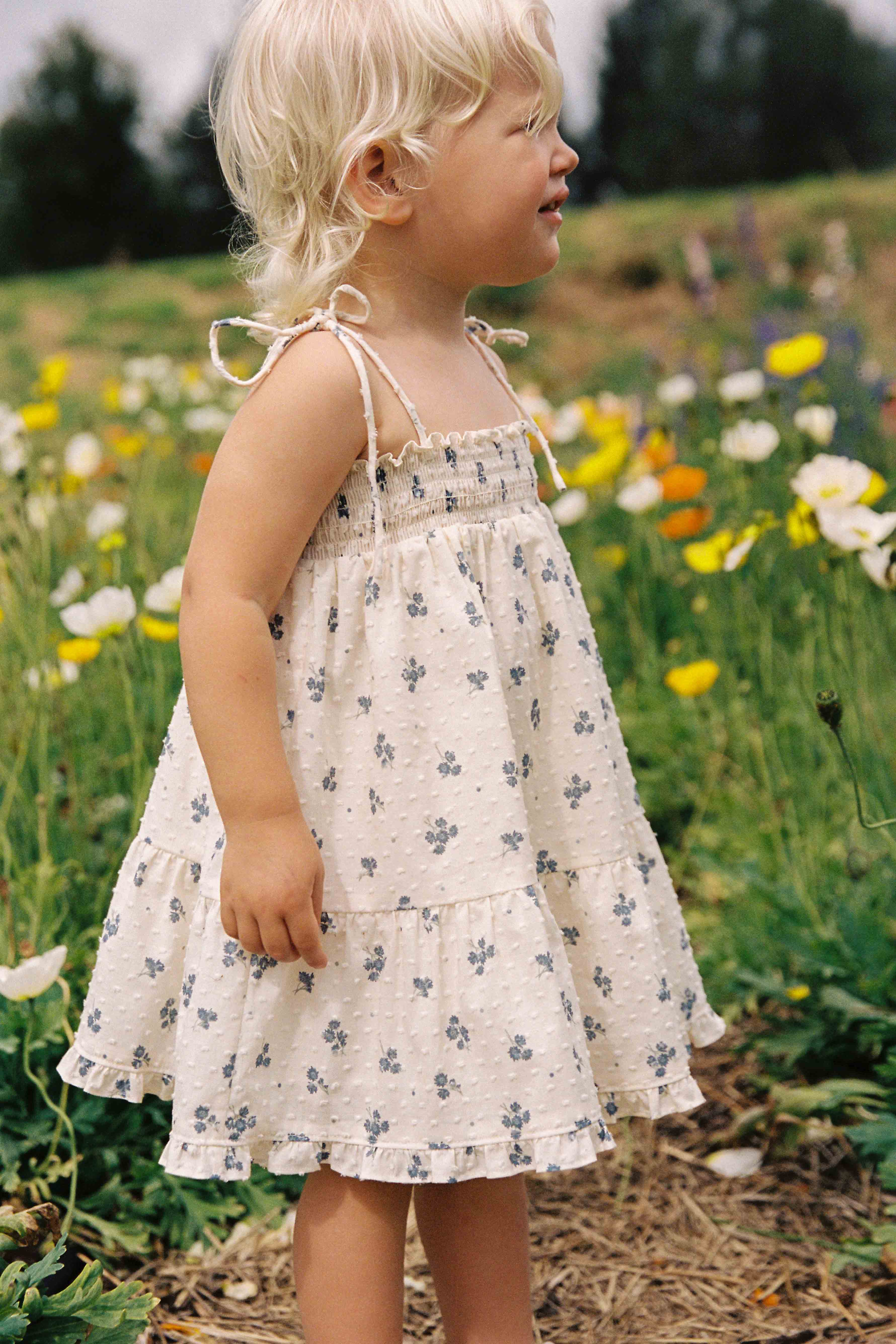 A young child with blonde hair stands in profile in a field of wildflowers, wearing JUNI JUNIOR's Posy Dress Ditsy Floral, a white sleeveless dress made of textured cotton dobby featuring small floral patterns and adjustable straps. The surrounding field is filled with diverse, colorful flowers and greenery.