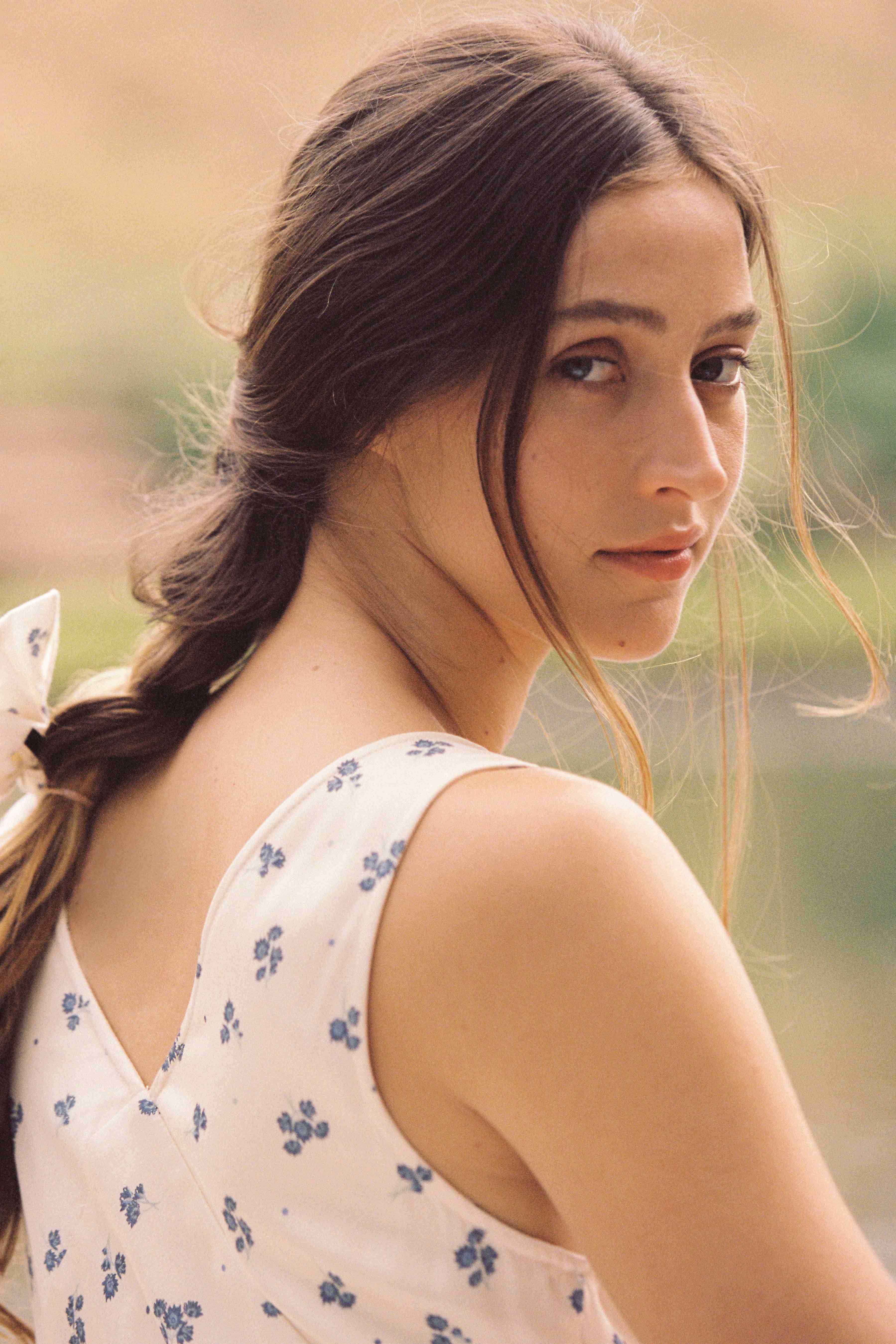 A young woman with long brown hair styled in a loose braid looks over her shoulder. Her hair is tied with a white ribbon. She is wearing the ethically made Isabella Midi Dress Ditsy Floral by JUNI, featuring a sleeveless design, blue floral patterns, and a delicate V neckline. The background is blurred, hinting at greenery and nature.