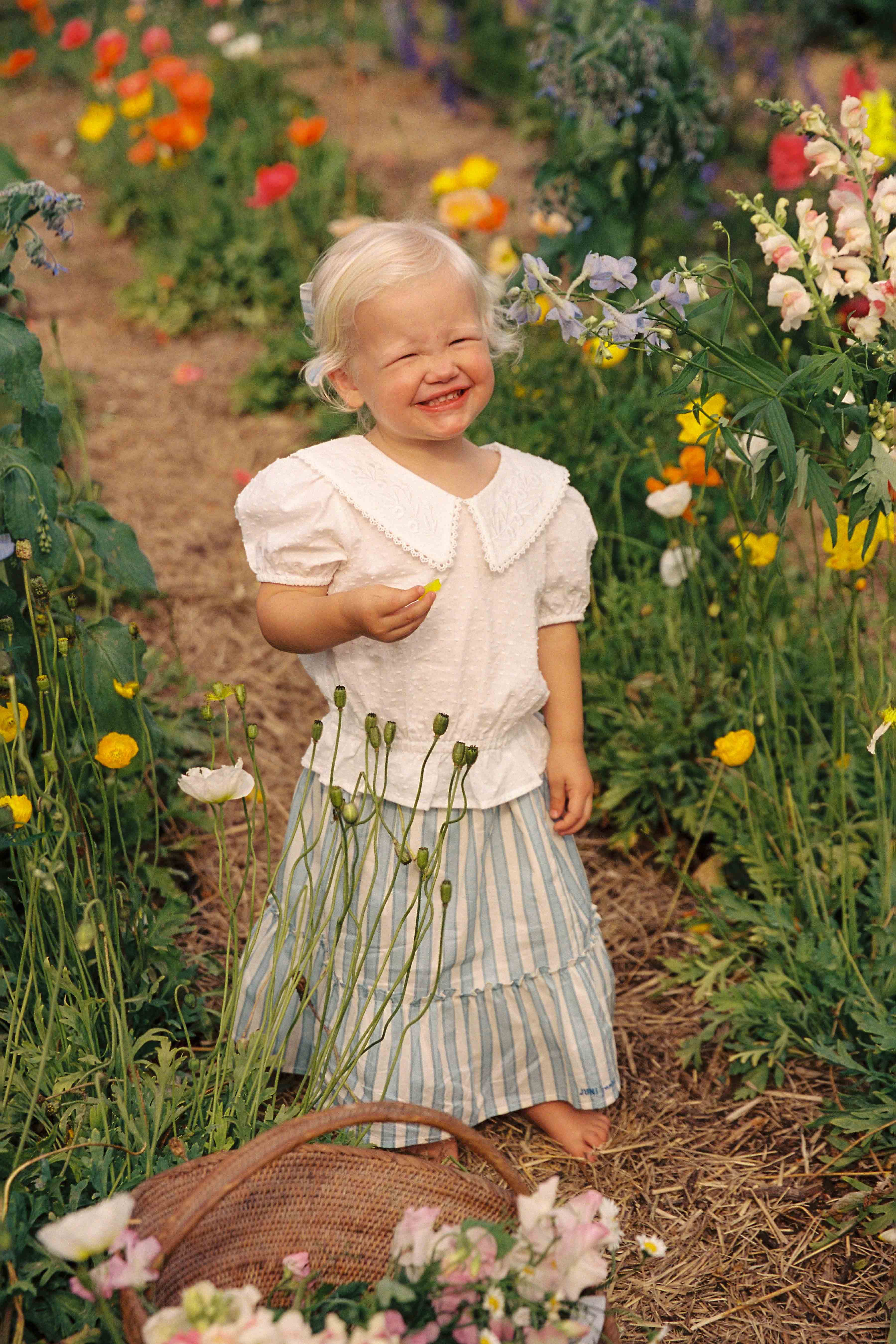 A young child with blonde hair stands in a flower garden, smiling with eyes closed. The child is wearing an ethically made, white cotton dobby Junior Celine Blouse with a Peter Pan collar from JUNI JUNIOR and a light blue striped skirt. Holding a small flower in one hand, they stand beside a wicker basket full of flowers.