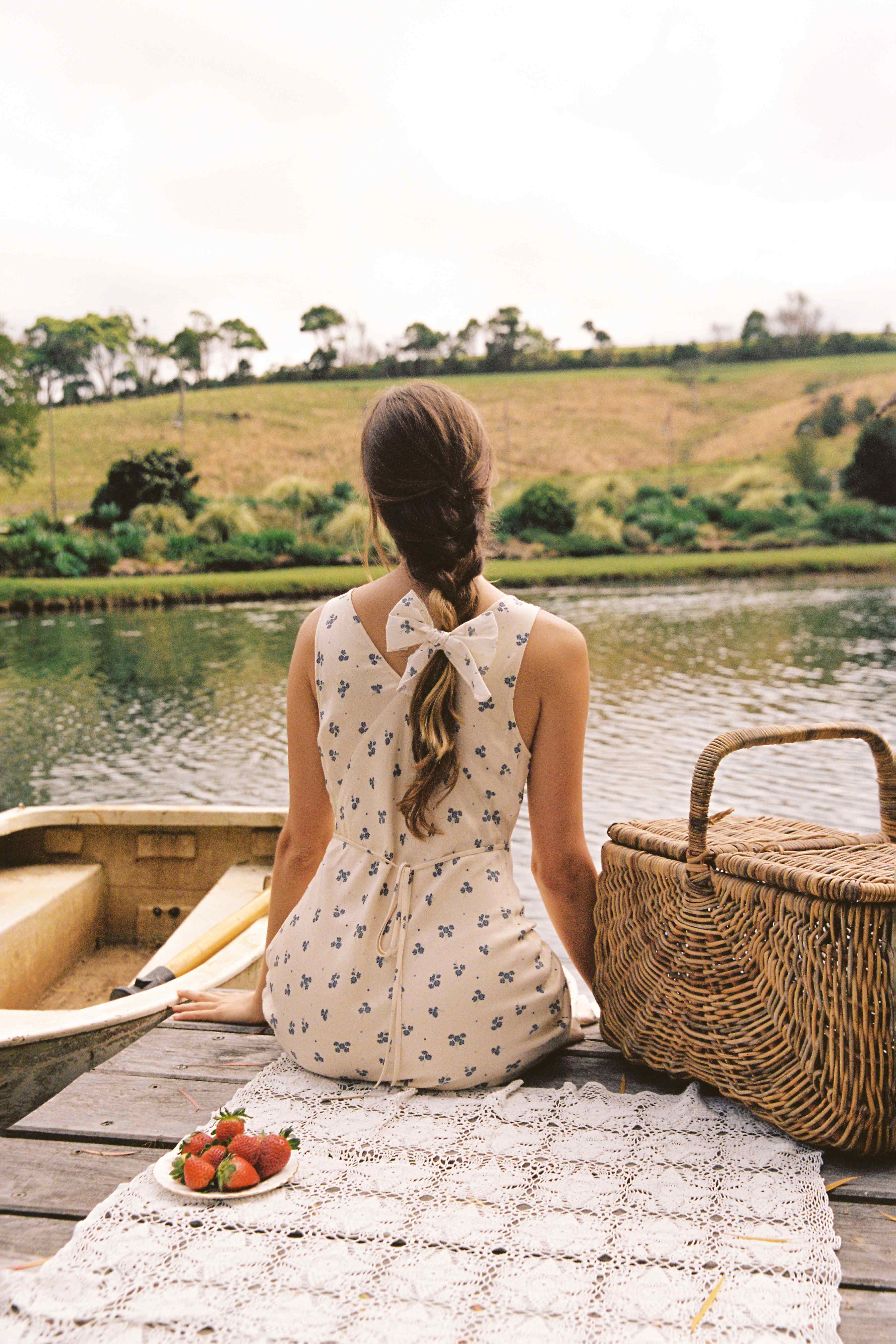 A woman with long braided hair, wearing the JUNI Isabella Midi Dress Ditsy Floral, sits on a dock by the water. She is next to a picnic basket and a lace blanket with a plate of strawberries. In the background is a scenic landscape with trees and rolling hills.