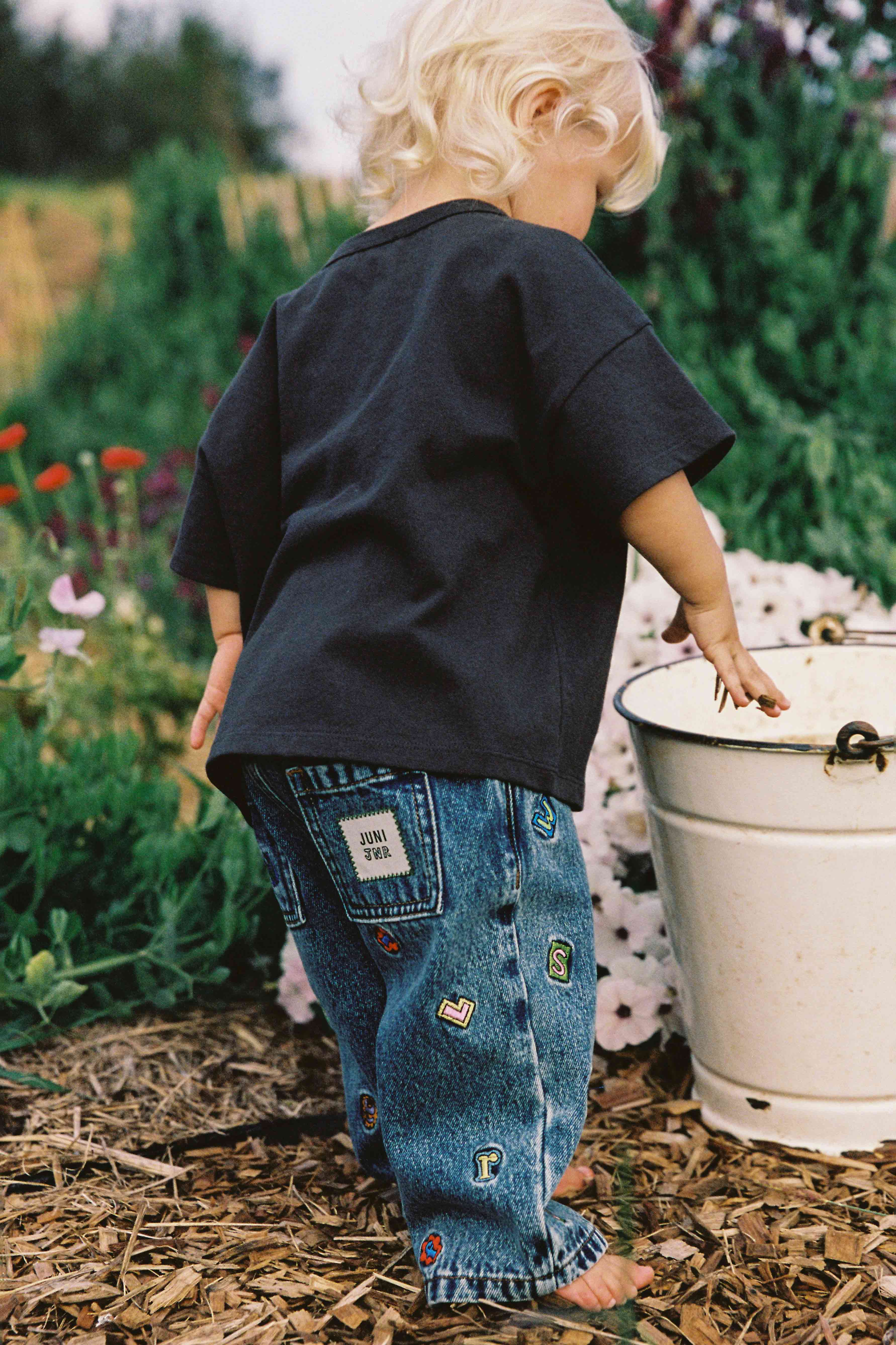 A small child with curly blonde hair, wearing a dark T-shirt and the Alphabet Denim Jean with colorful patches and an elastic waistband by JUNI JUNIOR, stands outdoors near a white metal bucket. The background features plants, flowers, and bark mulch on the ground.