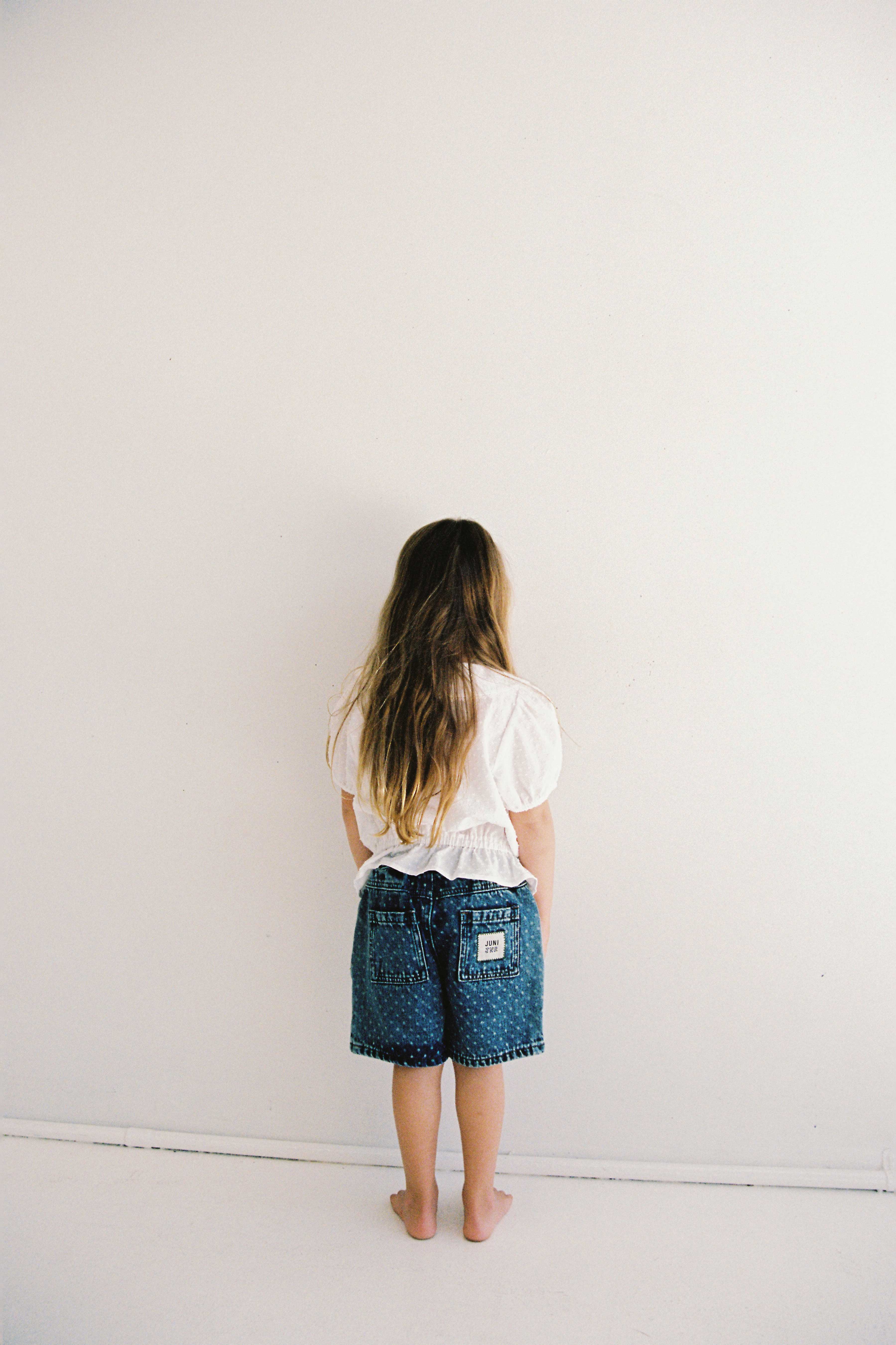 A child with long brown hair, wearing a white shirt and the Junior Bud Denim Short Spot by JUNI JUNIOR, stands barefoot facing a white wall. The setting appears to be minimalistic with a clean, empty space. The blue high-waist denim shorts feature an elastic waistband made from 100% cotton denim, ensuring both comfort and durability.