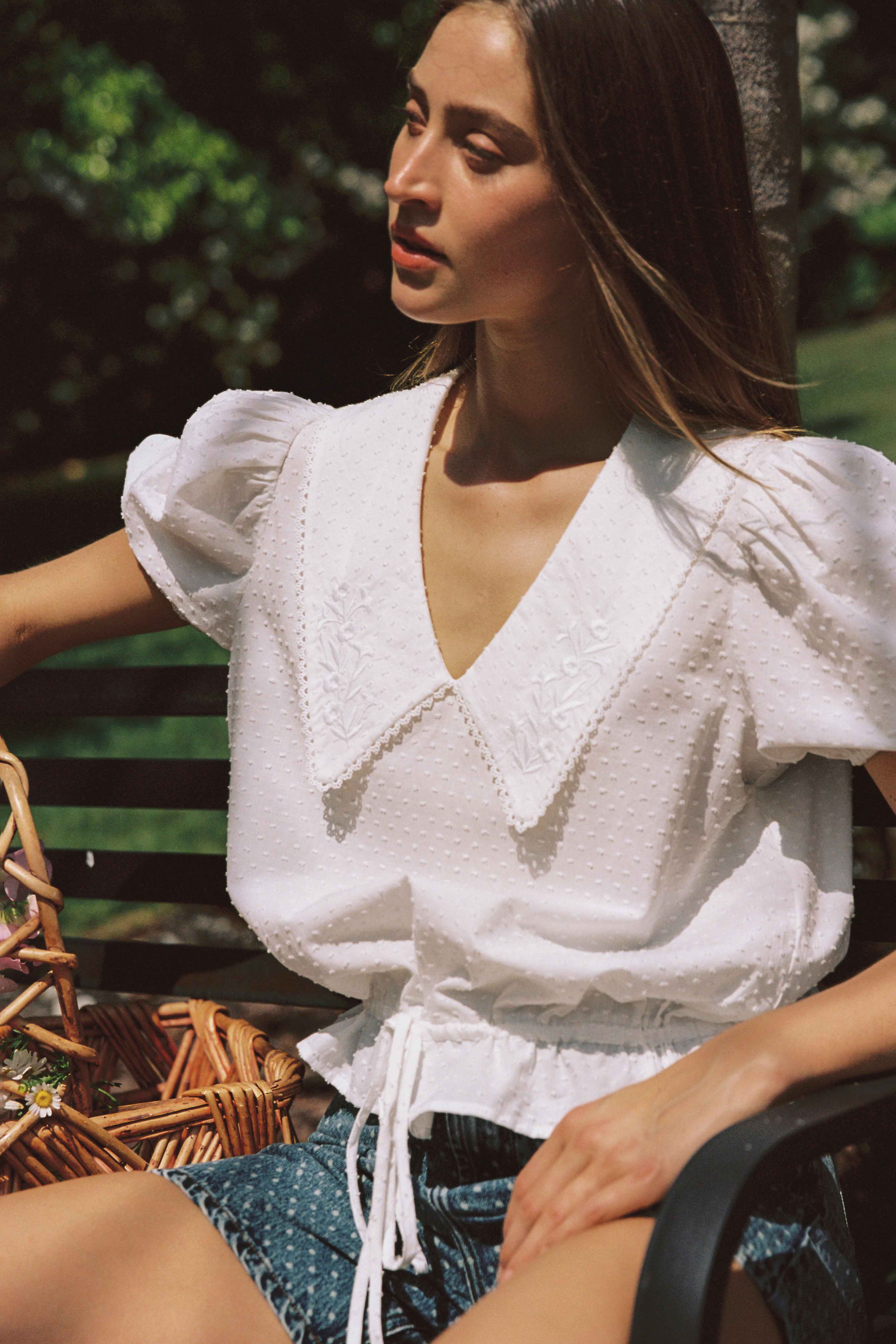 A woman with long hair sits on a park bench, wearing the Celine Short Sleeve Blouse by JUNI, featuring intricate embroidery and paired with blue shorts made of soft dobby cotton. Next to her is a wicker basket. Sunlight filters through the trees, casting a warm glow on her face.