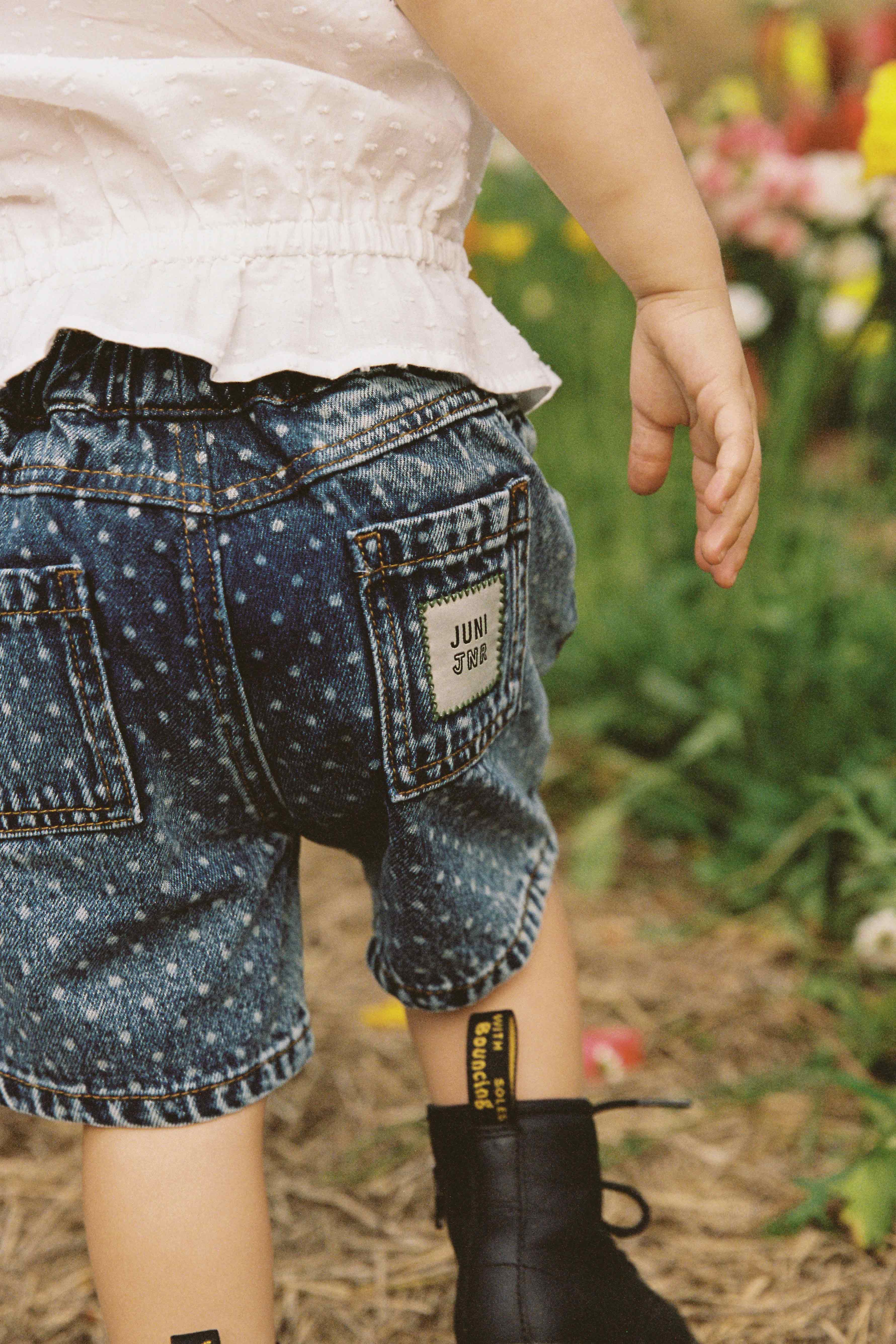 A small child stands on a grassy surface, dressed in a white blouse, black boots, and JUNI JUNIOR's Junior Bud Denim Short Spot – blue high-waist denim shorts made from 100% cotton. The back pocket of the shorts features a text patch. The background is blurred with hints of flowers and greenery visible. The child's face is not shown.