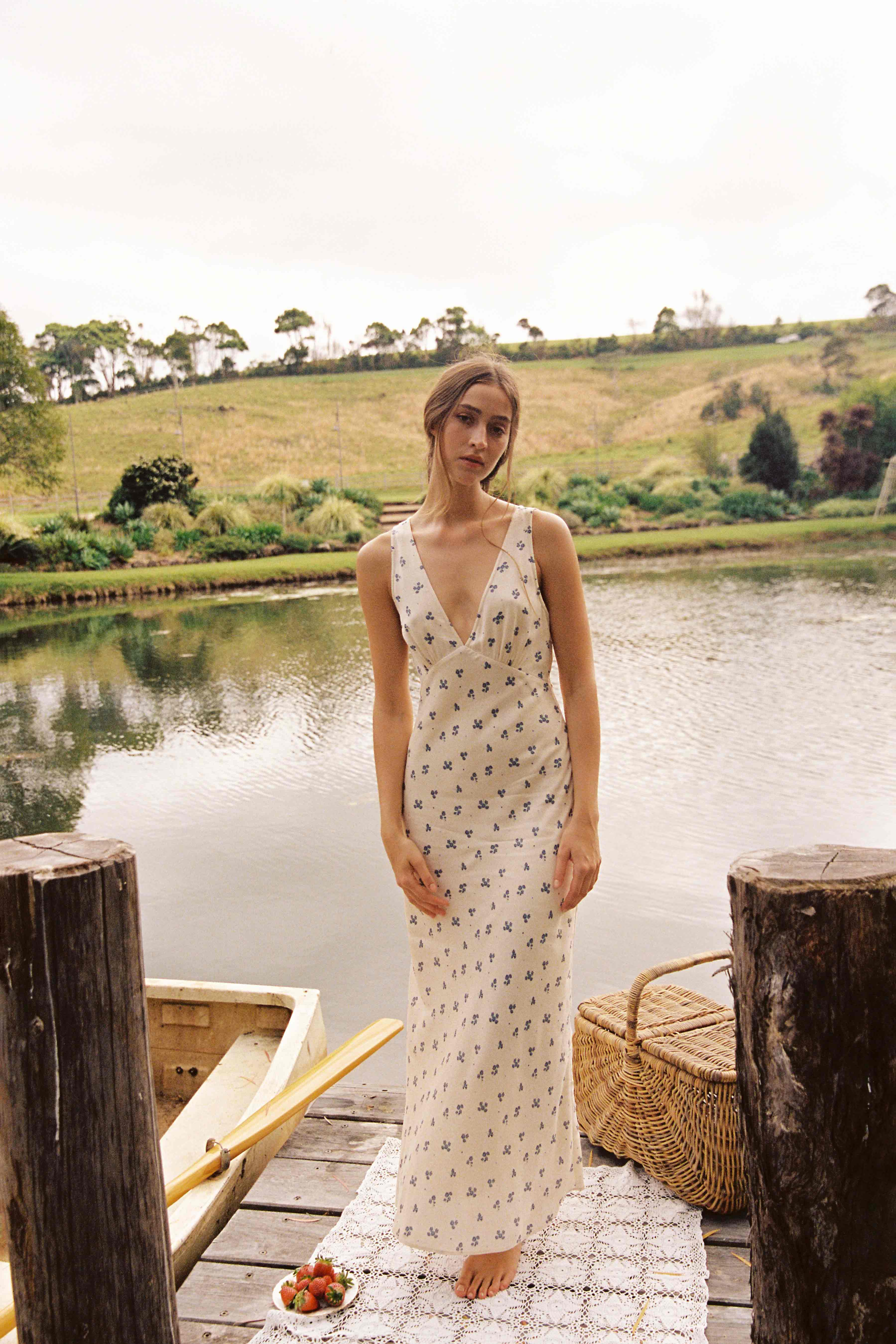 A woman stands on a wooden dock near a serene lake, with grassy hills and trees in the background. She wears an ethically made Isabella Midi Dress Ditsy Floral from JUNI featuring a bias cut and a v neckline. A boat is docked beside the pier, and a basket with strawberries is placed on the dock.