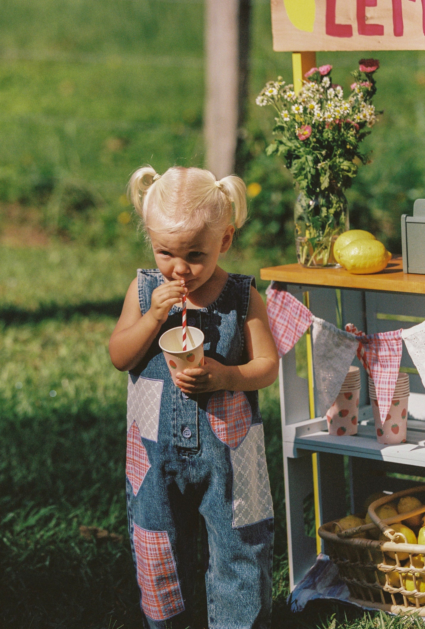 A little child with blonde pigtails stands on grass, wearing JUNI JNR Charlie Overalls, sipping through a straw. In the background is a lemonade stand decorated with flowers, lemons, and a basket.