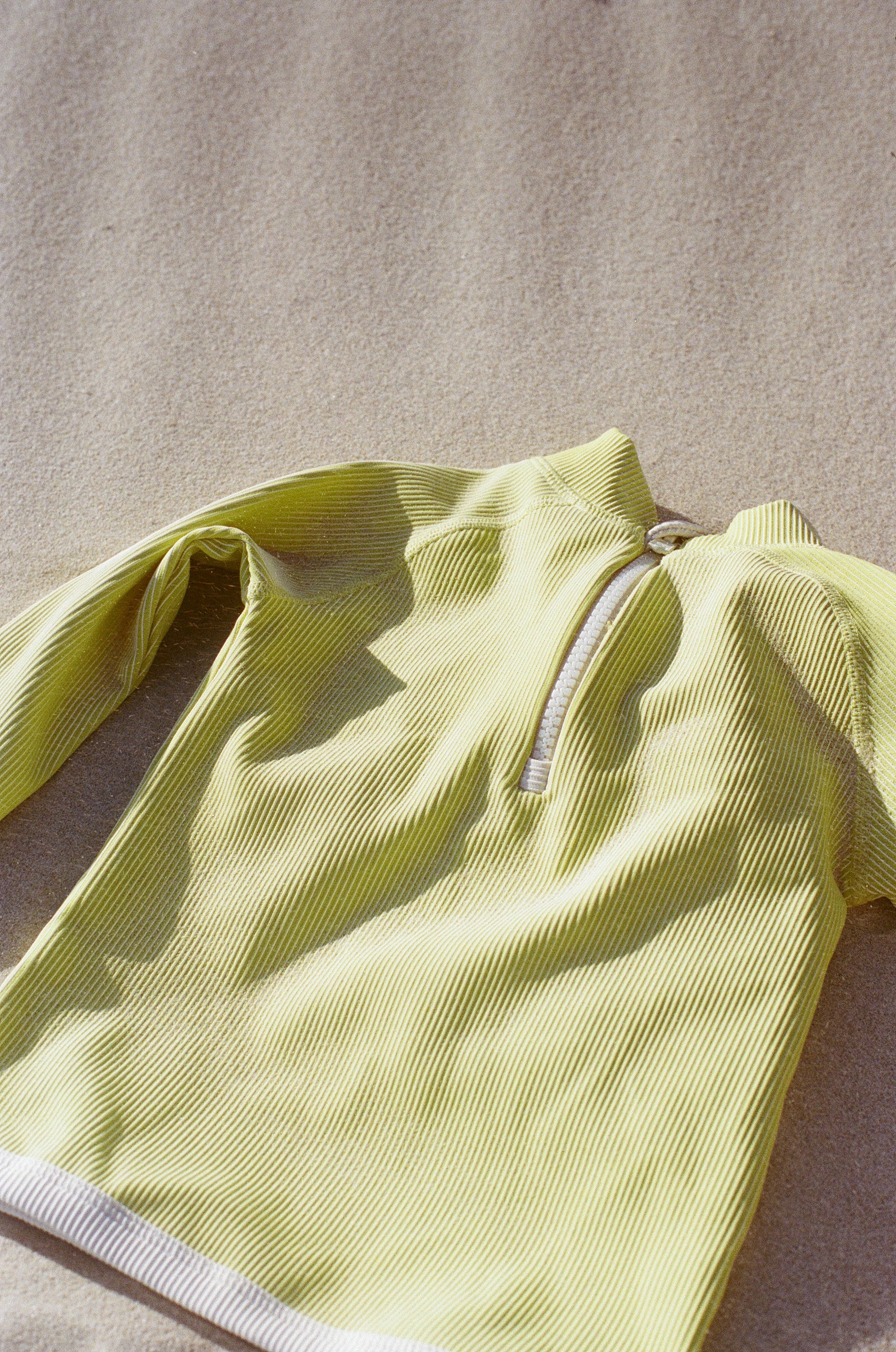 The PRE-ORDER Mini Stripe Rashie Citrus by ZULU & ZEPHYR, a light yellow long-sleeve pullover with a half-zip front crafted from recycled materials, lies flat on a beige surface, with soft shadows highlighting its textures and folds.