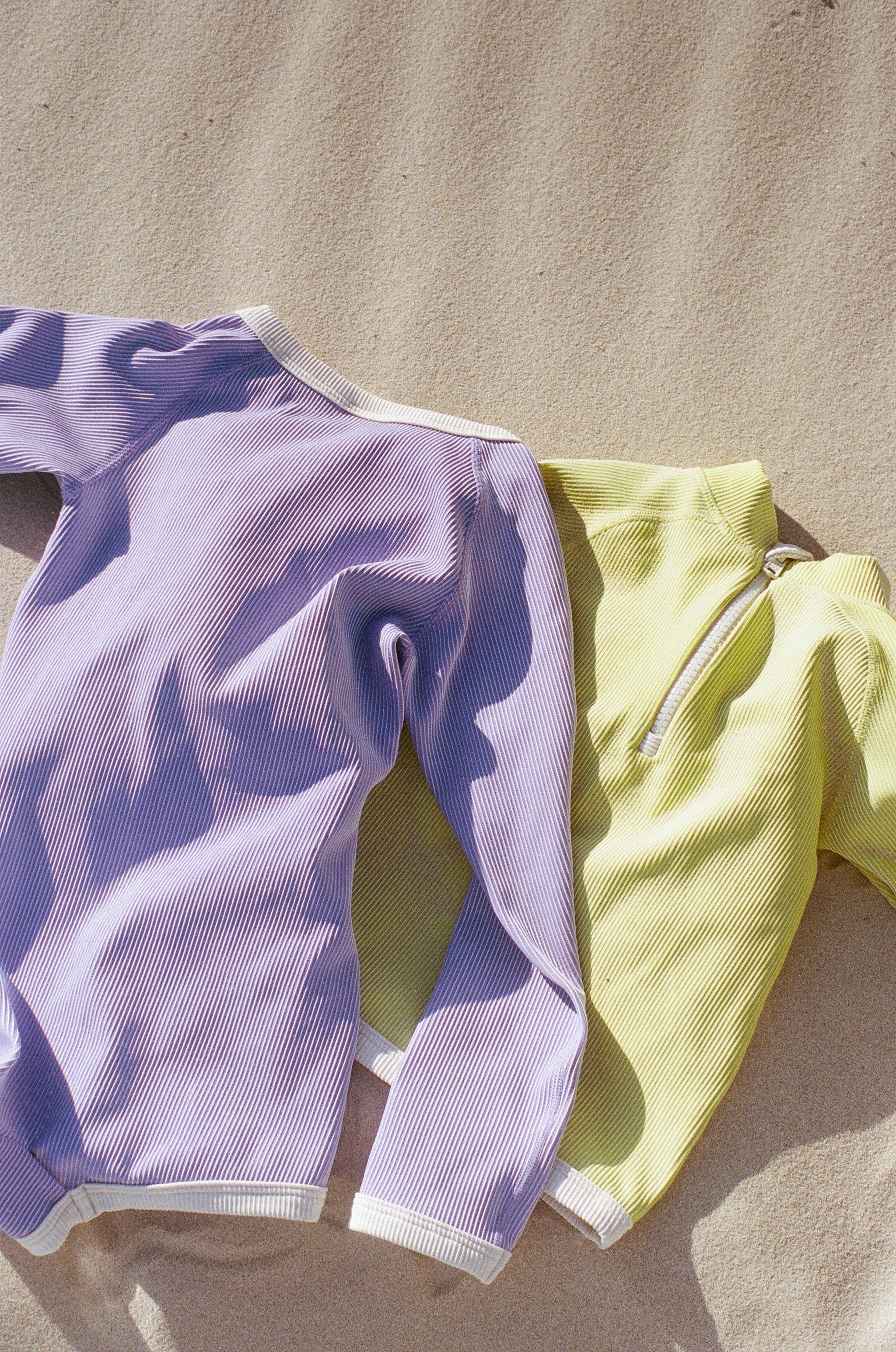 Two long-sleeved ribbed shirts lie on the sandy ground. One shirt is lavender with white edges, and the other is a pastel yellow PRE-ORDER Mini Stripe Rib Rashie Onesie Citrus from ZULU & ZEPHYR, featuring a white zipper and edges. Made from recycled Lycra, they offer UPF 50+ protection. Sunlight casts soft shadows on the fabric, highlighting the shirts' texture.