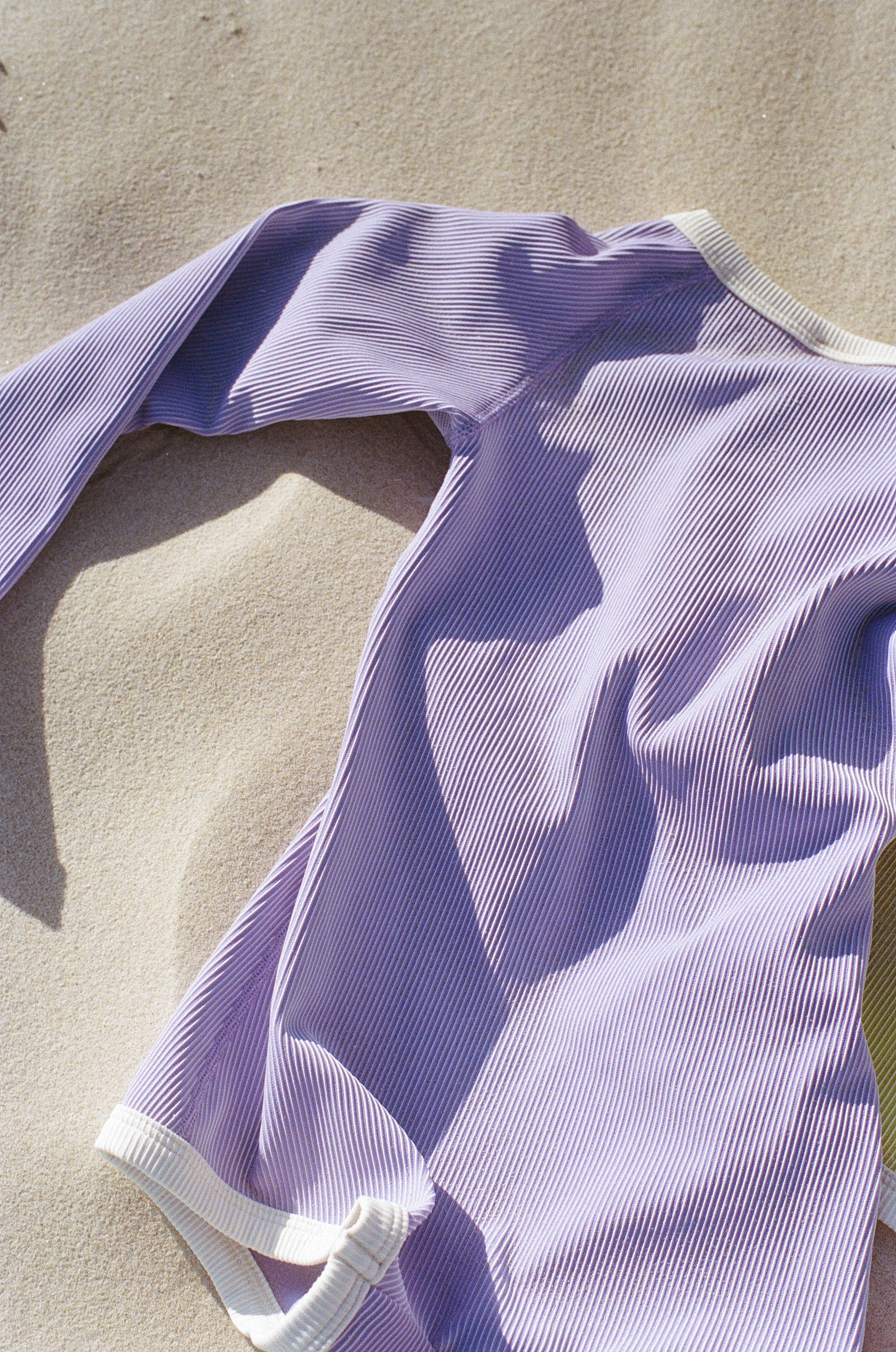 A PRE-ORDER Mini Stripe Rib Rashie Onesie in Grape by ZULU & ZEPHYR, featuring long sleeves and white trim, is laid out on a sandy beach with sunlight casting shadows across its pastel purple fabric made from recycled materials.