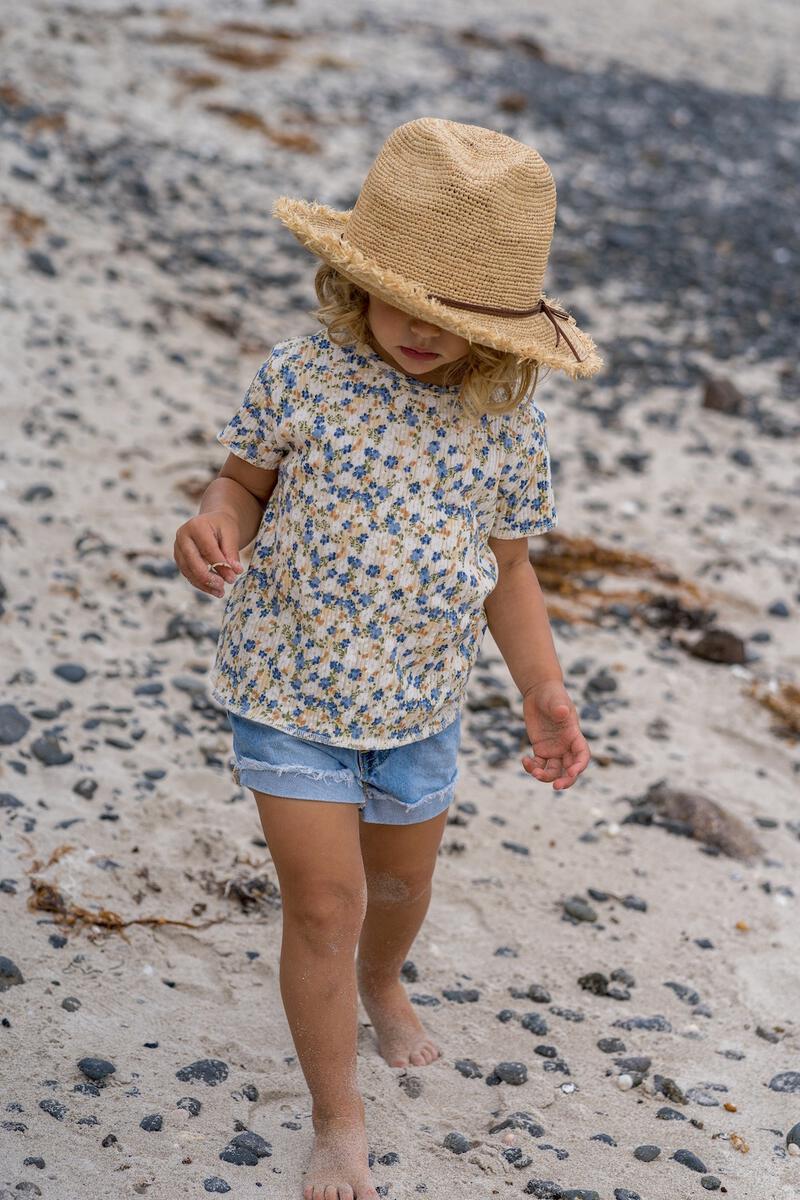 A young blond child wears the ACORN KIDS Acorn Kids - Coco Straw Hat with a floral shirt and denim shorts, walking barefoot on a rocky, sandy beach.