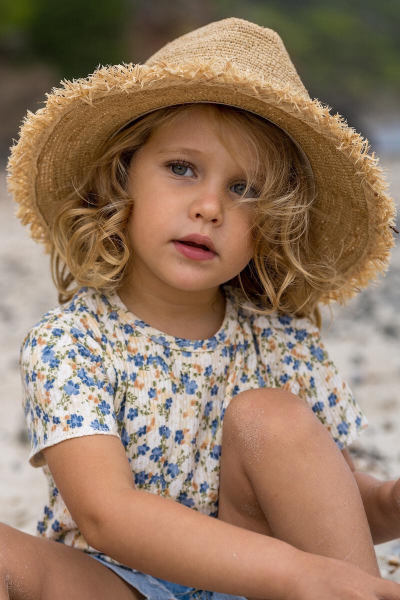 A young child in the ACORN KIDS Acorn Kids - Coco Straw Hat sits on the sand, looking thoughtfully at the camera, enjoying sun protection.