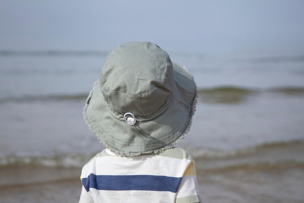 A young child wearing the Acorn Kids - Frayed Bucket Hat Khaki by ACORN KIDS and a striped shirt stands on the shore, gazing at the sea with gentle waves beneath a clear sky.