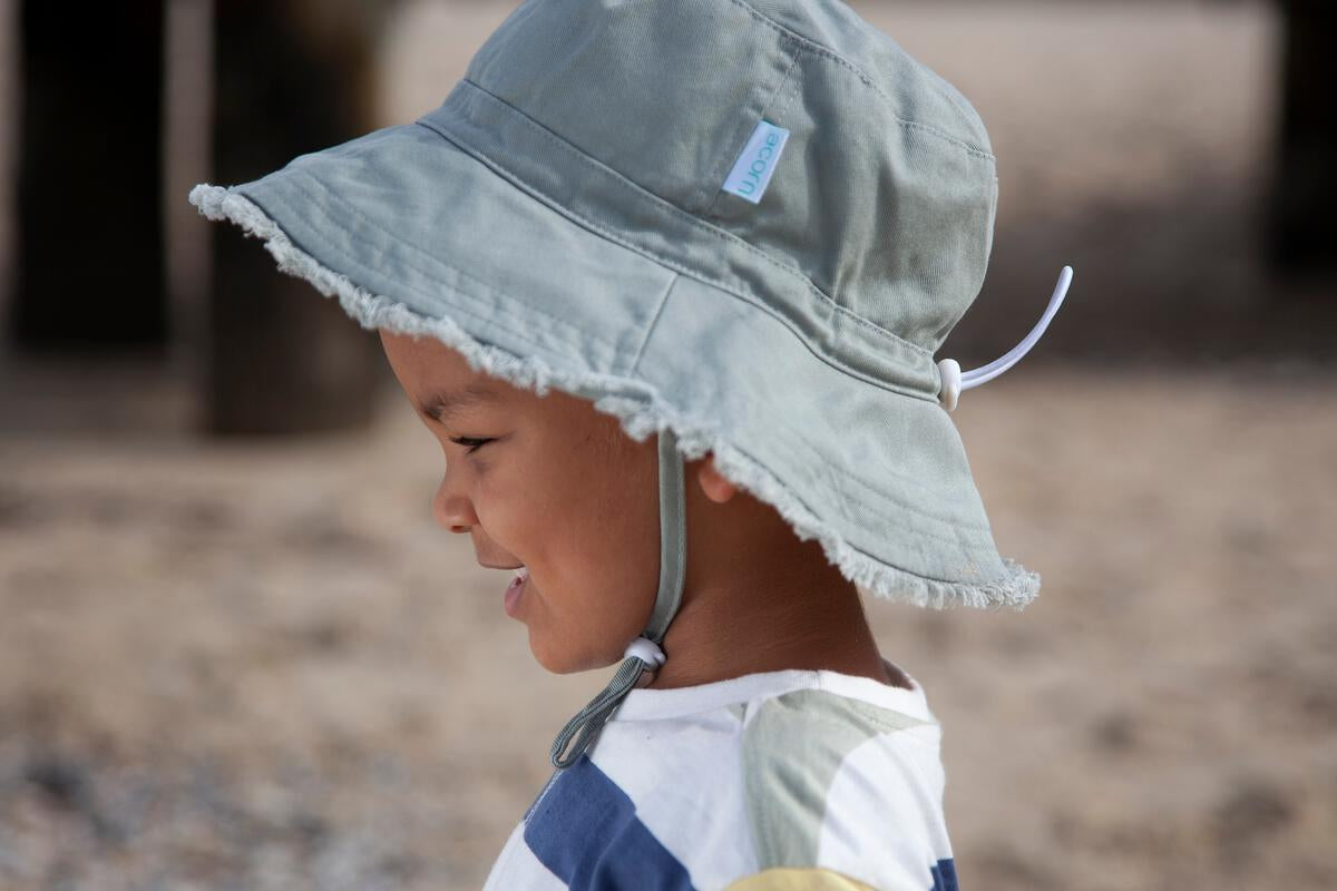A young child in the ACORN KIDS Acorn Kids - Frayed Bucket Hat Khaki smiles outdoors on a sandy beach, also wearing a striped shirt.