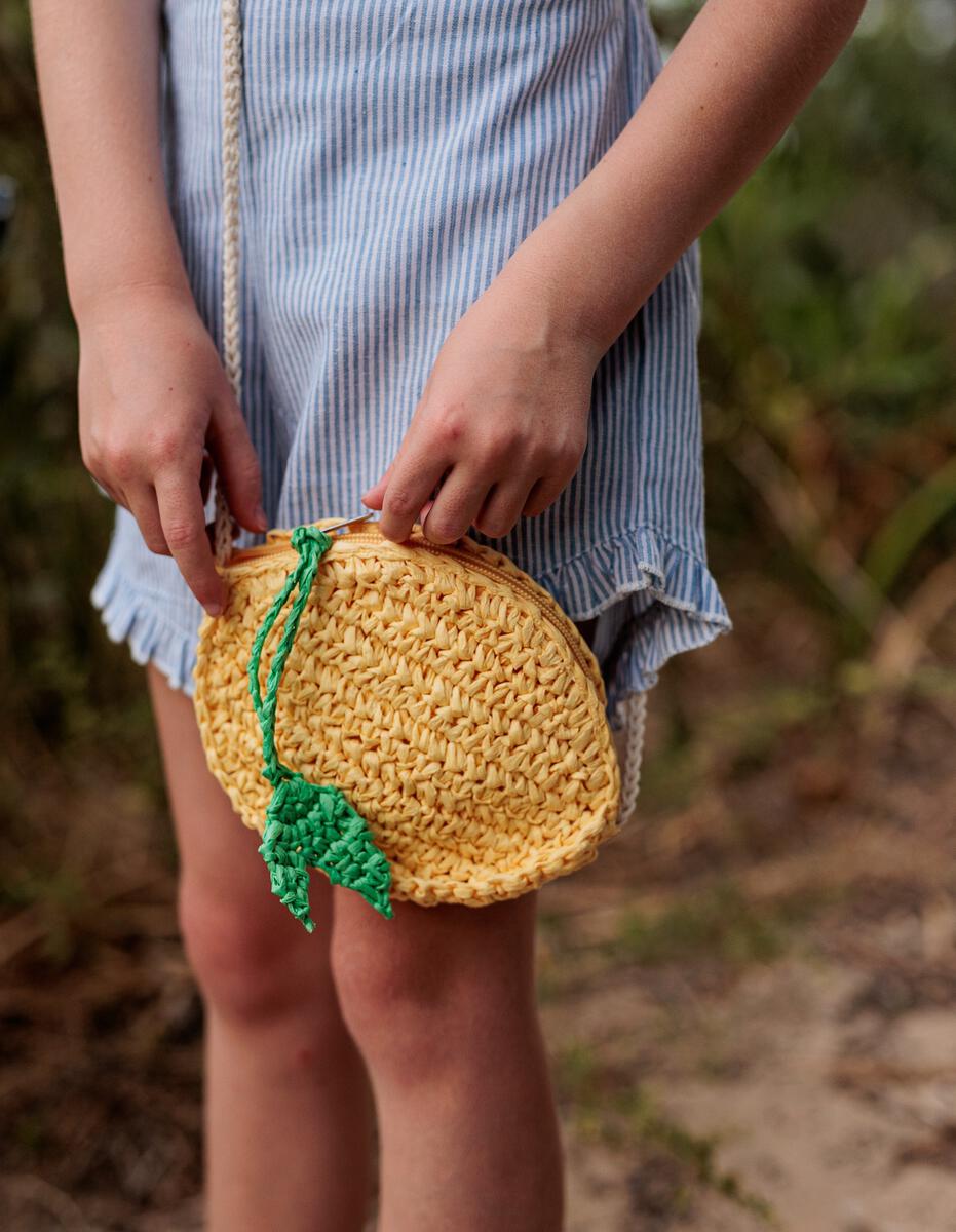 A child in a blue and white striped outfit holds the Acorn Kids - Ravello Lemon Straw Bag by ACORN KIDS, featuring a cute lemon design with a green leaf, outdoors.
