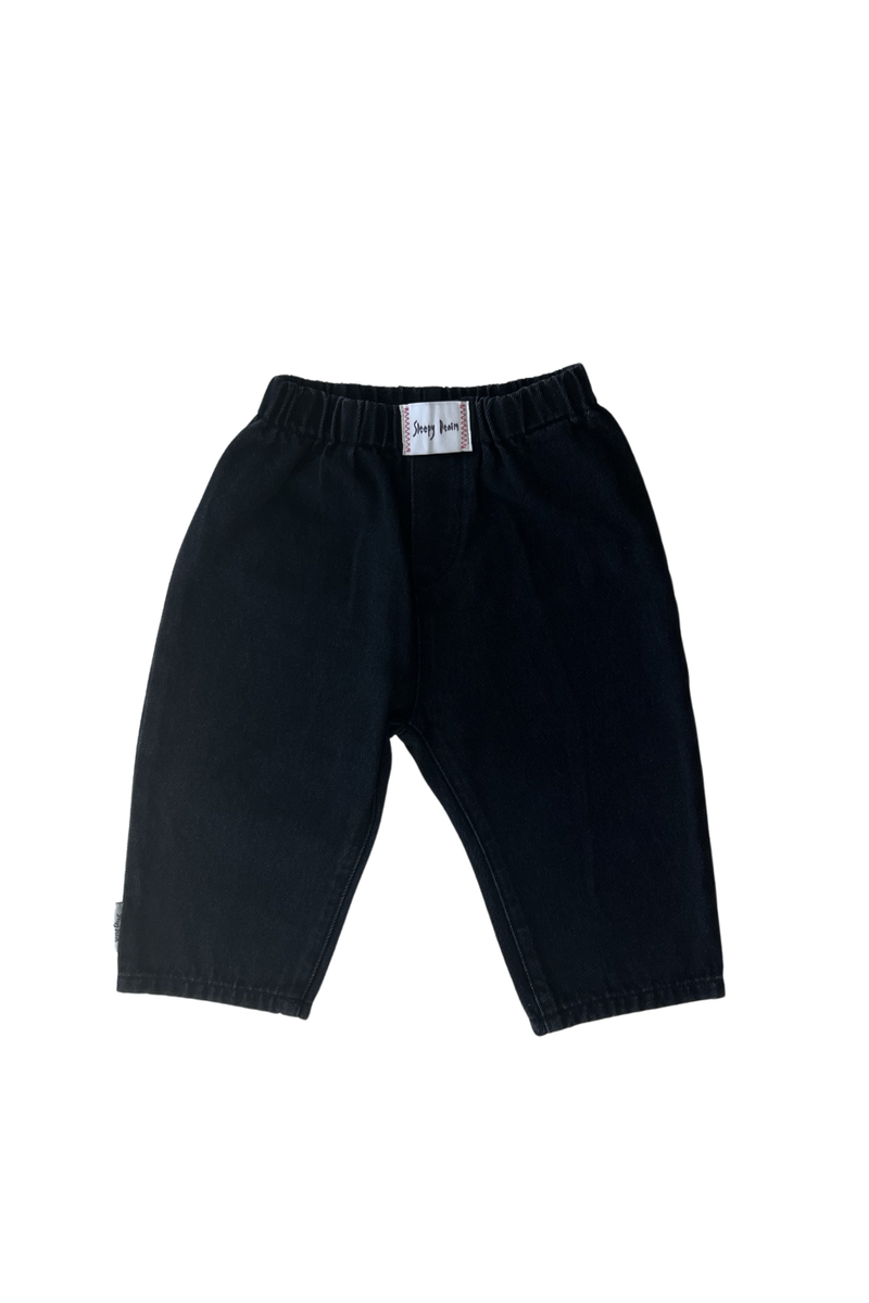 Introducing the Raver Jean Black Denim by SLEEPY DENIM: These black, knee-length children's denim shorts feature an elastic waistband for comfort. The minimalist design includes no visible pockets or patterns, and there's a small white tag with text at the center of the waistband. Made from organic cotton, they offer ultimate comfort and durability.