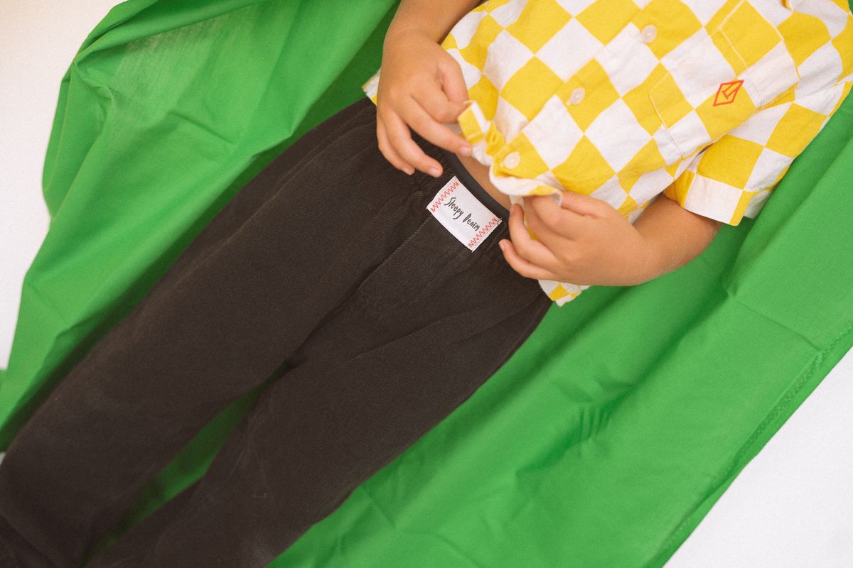 A child dressed in a yellow and white checkered shirt adjusts the waistband of their dark children's denim pants, which sport a visible SLEEPY DENIM label for the Raver Jean Black Denim. A bright green cloth drapes in the background, capturing the image from shoulders to knees.