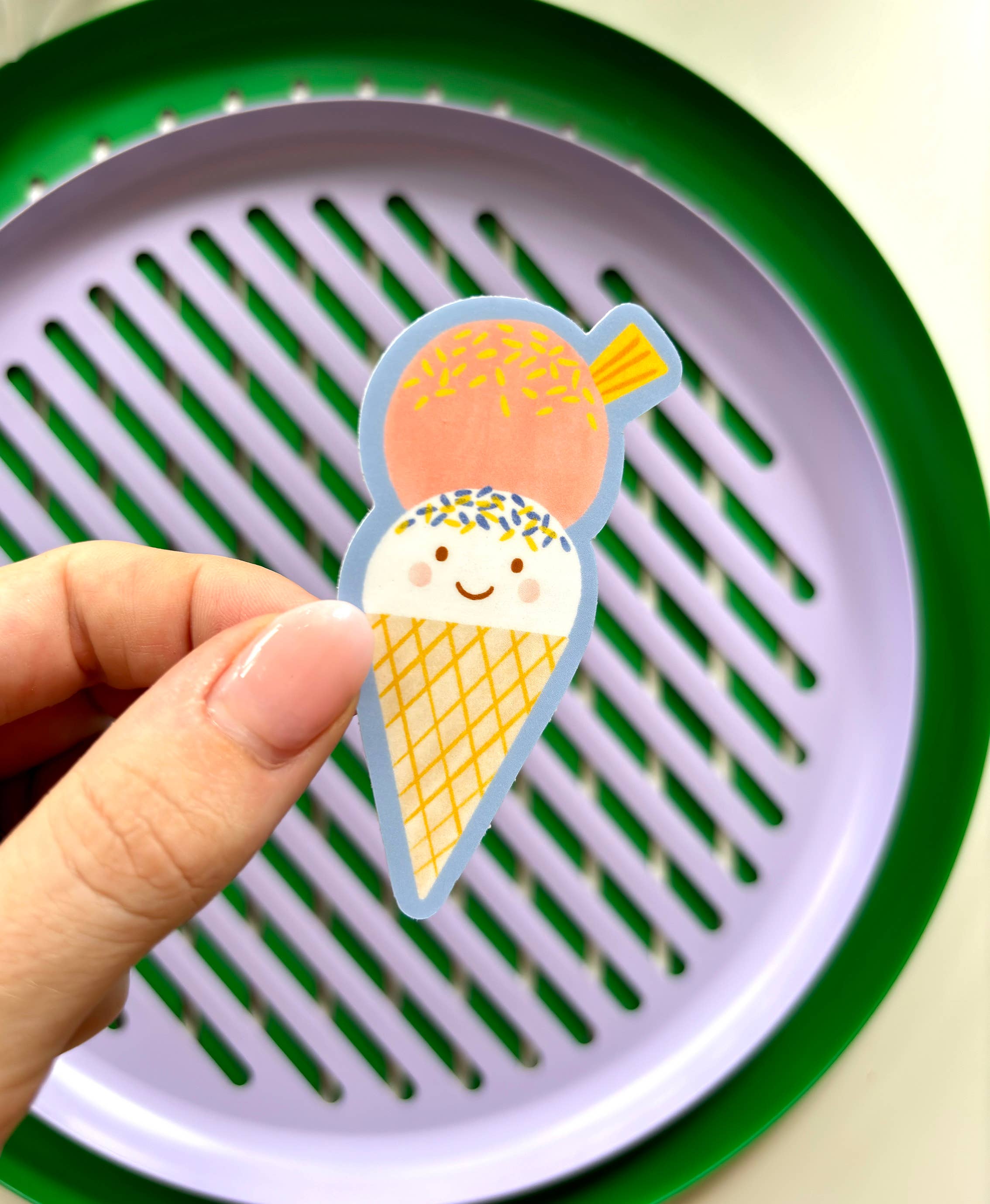 A hand holding the "Ice Cream Sticker" by LAUREN SISSONS STUDIO, featuring a cute, high-quality vinyl sticker of a two-scoop ice cream cone with smiling faces. The top scoop is pink with sprinkles, and the bottom scoop is white with blue and yellow sprinkles. The background consists of a purple and green plate, printed on UV-coated matte vinyl for durability.