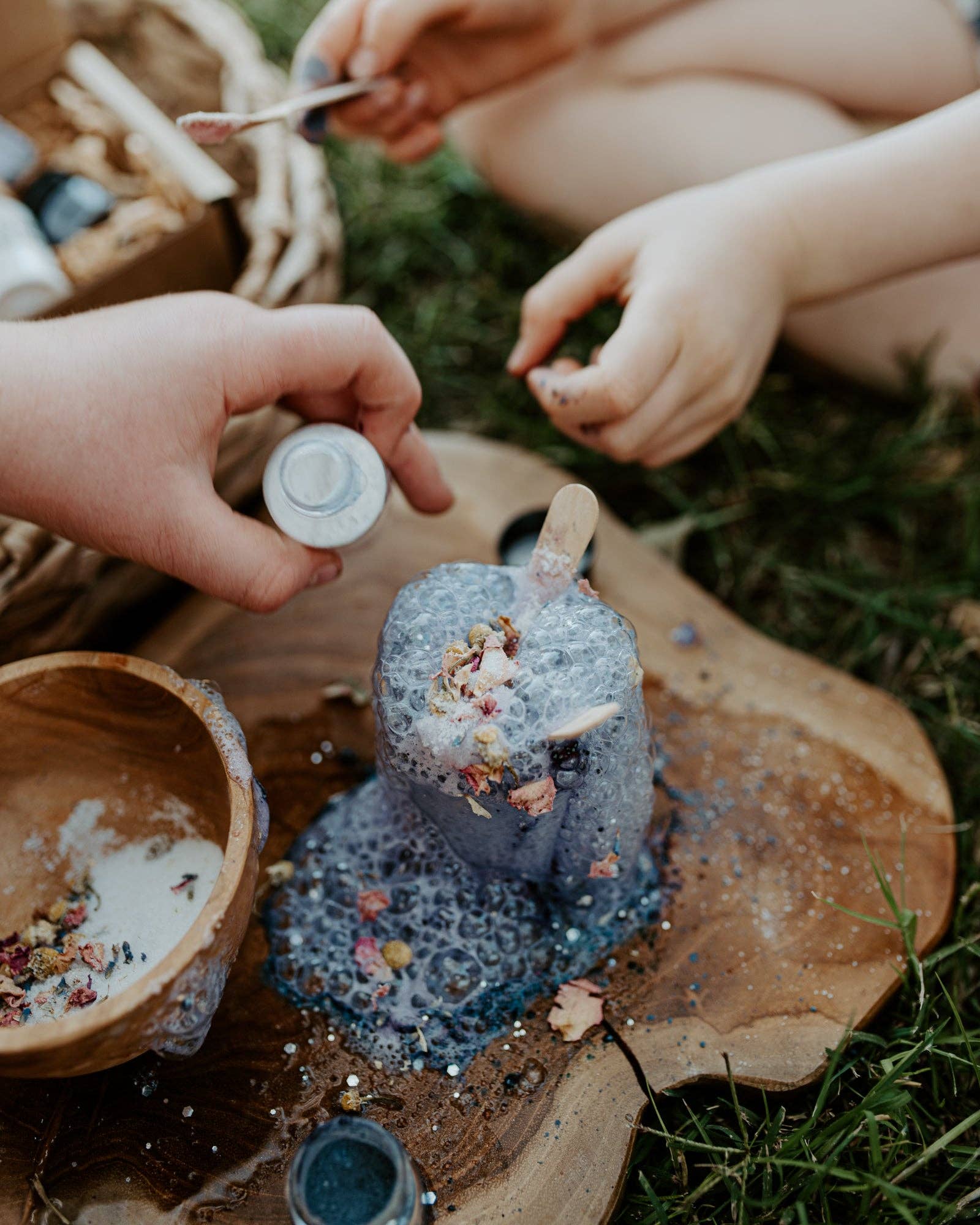 Two pairs of hands, possibly children, engage in a creative outdoor activity, adding glitter and small decorative items to a soapy bubble mixture on a wooden board. Various small containers and bowls with craft supplies are scattered around, resembling the enchanting setup of THE LITTLE POTION CO's MINI Colour Mood Potion Kit (Colour Changing Kit).