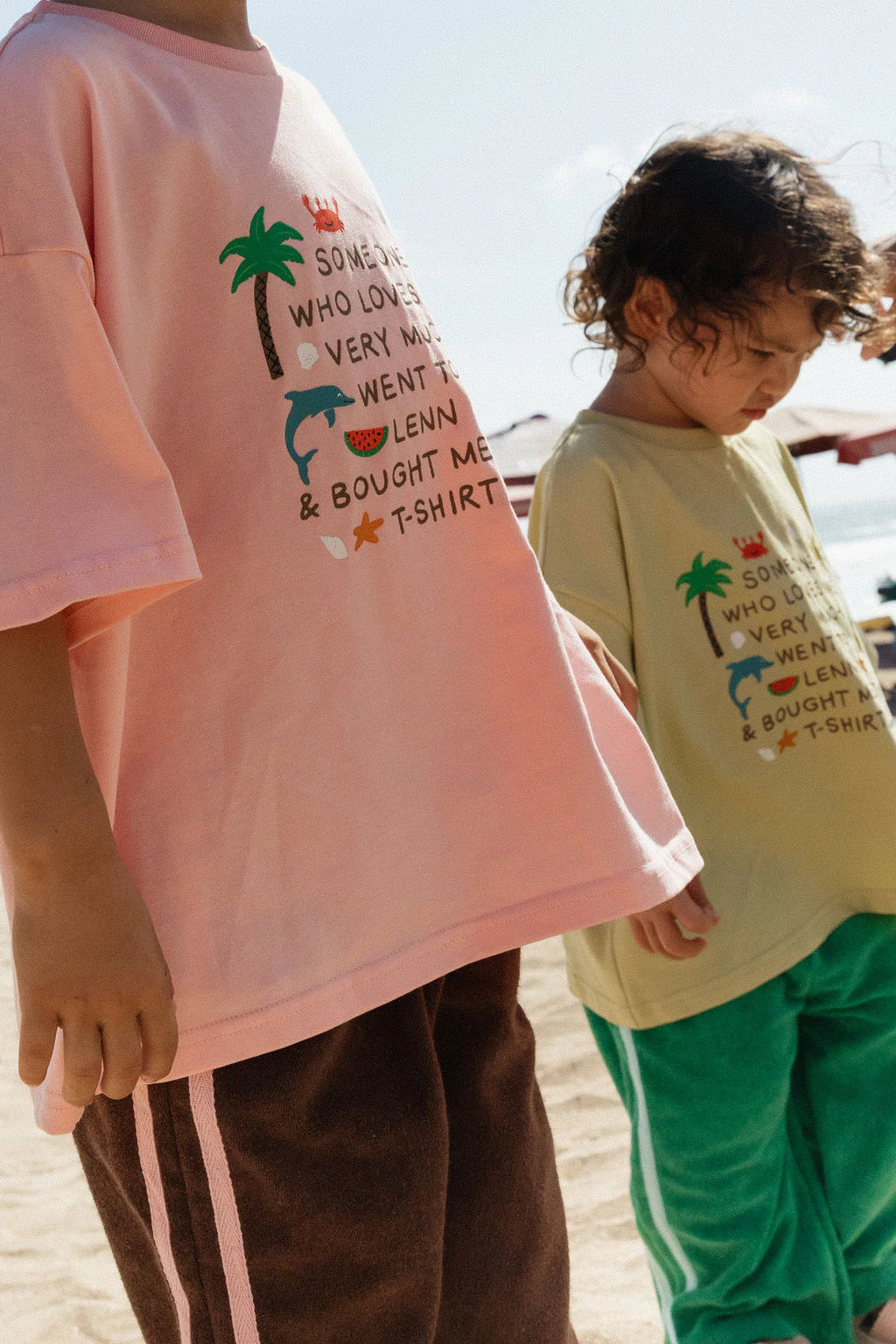Two kids stand on a sunny beach, each wearing LENN LABEL's Lenn Tee Blush—100% cotton, relaxed fit tees with playful graphics—one in blush pink with brown shorts, the other in yellow and green pants under a clear sky.