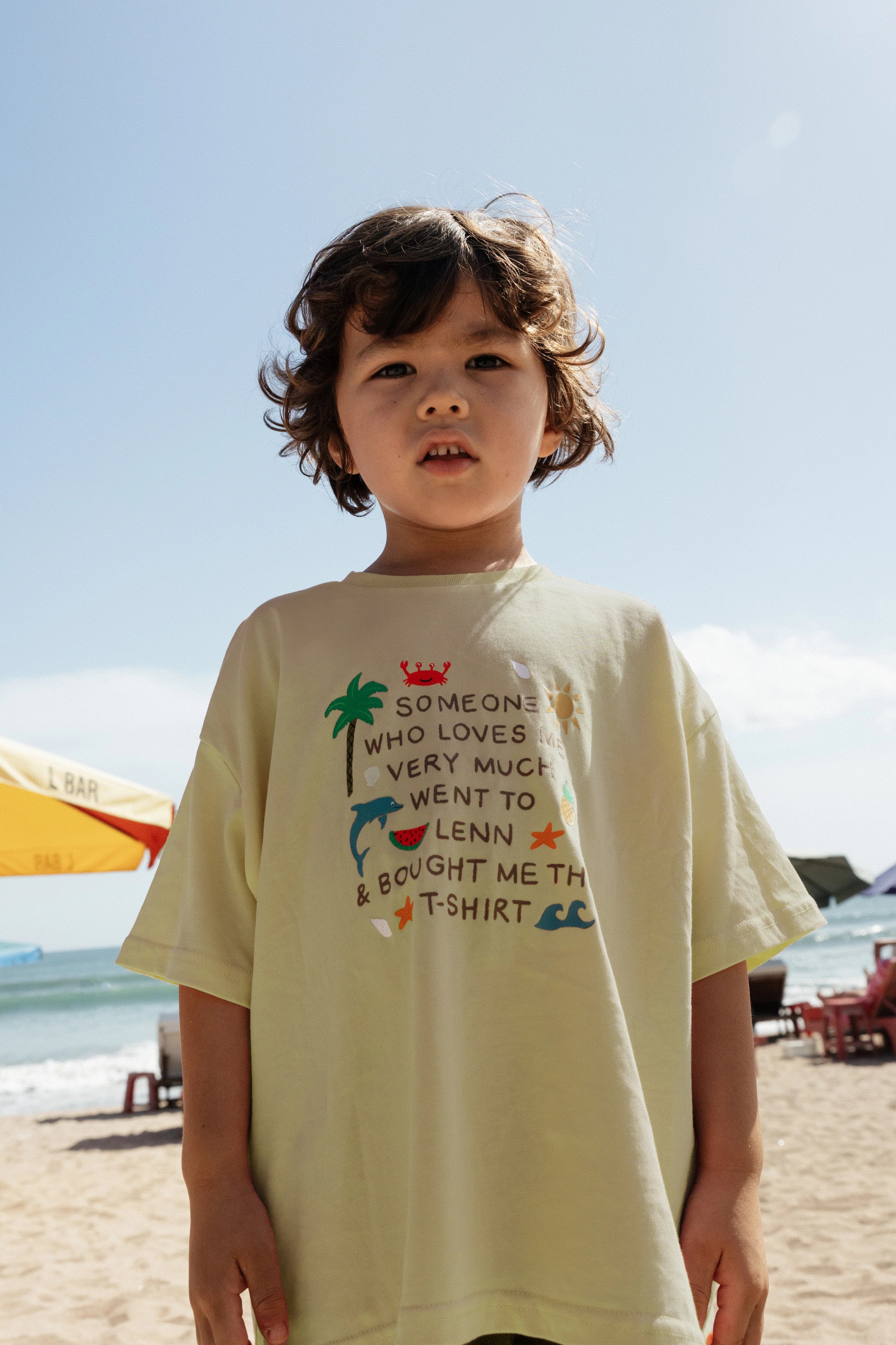 A young child with wavy hair stands on a sunny beach, wearing the LENN LABEL Lenn Tee Lime—a 100% cotton kids tee. Playful illustrations and text cover the lime shirt, as beach umbrellas and lounge chairs dot the shoreline in the background.