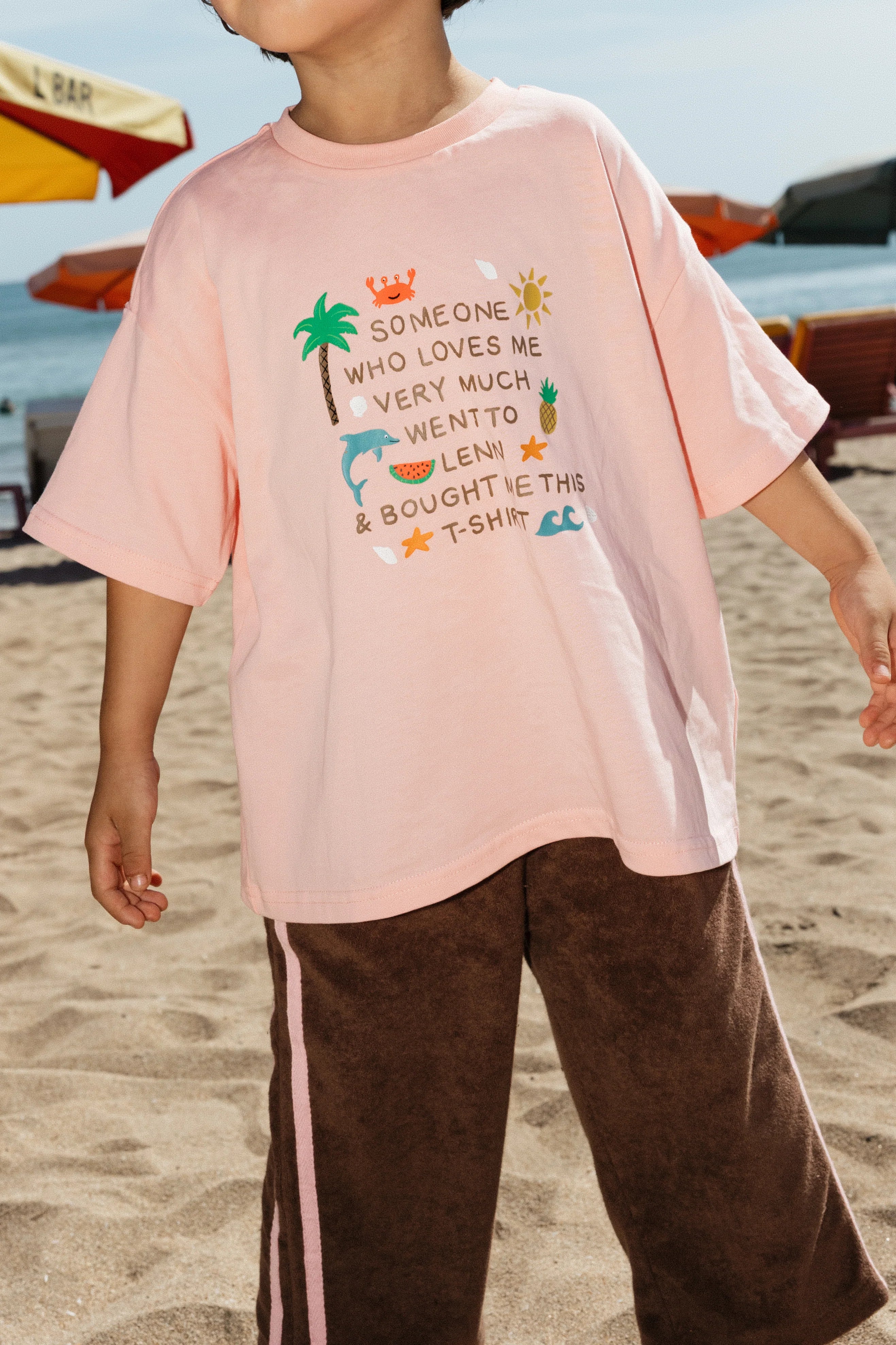 A child stands on the beach in the Lenn Label - Lenn Tee Blush by LENN LABEL, a relaxed fit, 100% cotton pink T-shirt with colorful graphics and the message, “Someone who loves me very much went to Leni & bought me this T-shirt.”.