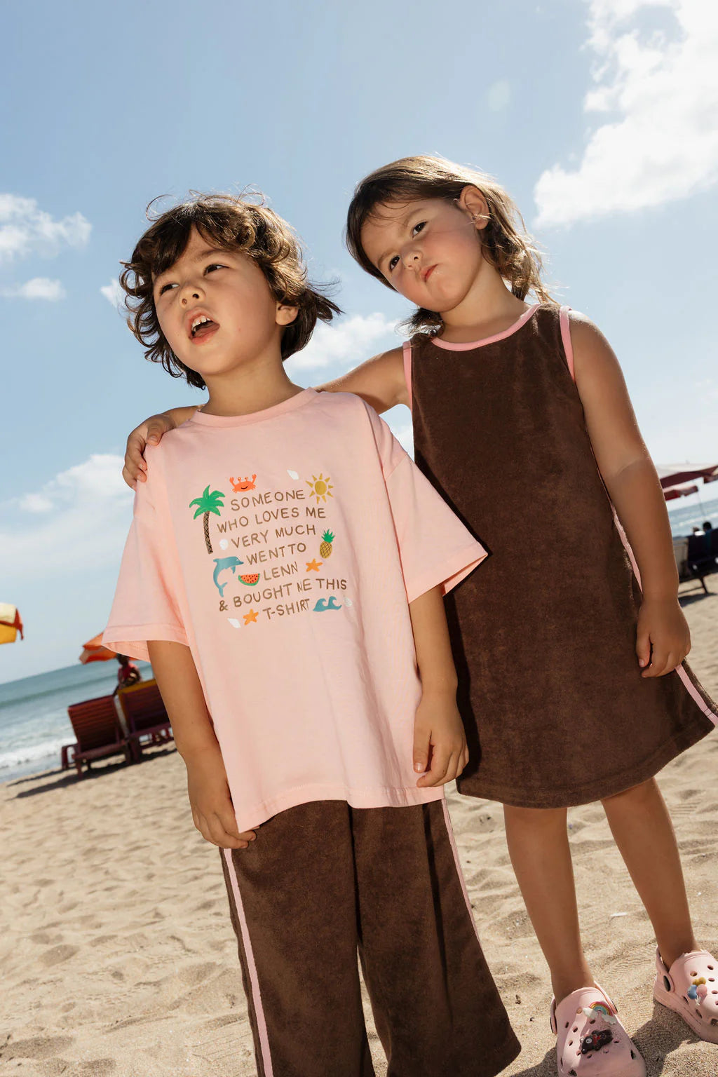 Two kids stand on a sunny beach. The boy wears the LENN LABEL - Lenn Tee Blush with brown shorts, and the girl wears a brown dress and pink clogs, her arm around his shoulder. Beach chairs and umbrellas are seen behind them.