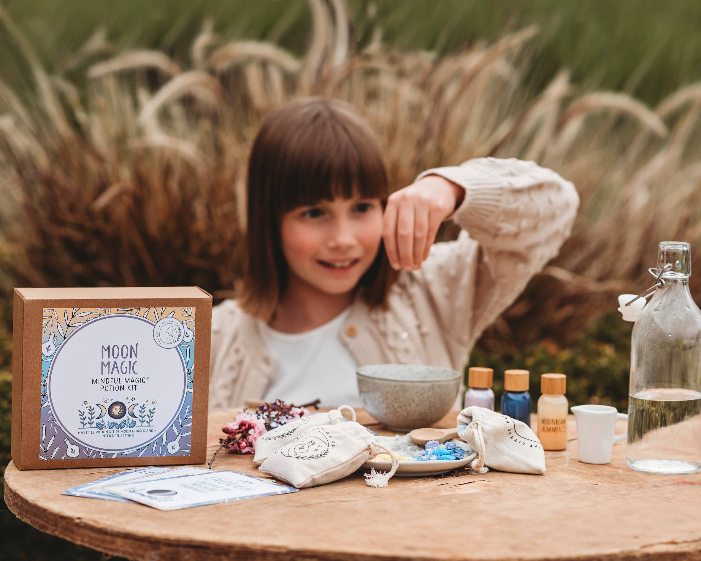 A young girl smiles as she sprinkles ingredients into a bowl on a table outdoors. The table is filled with items from THE LITTLE POTION CO's "Moon Magic MINDFUL Magic Potion Kit," including small bottles, pouches, and a box adorned with whimsical designs. This mindfulness activity unfolds under the gentle gaze of the moon phases, with grassy plants in the background.