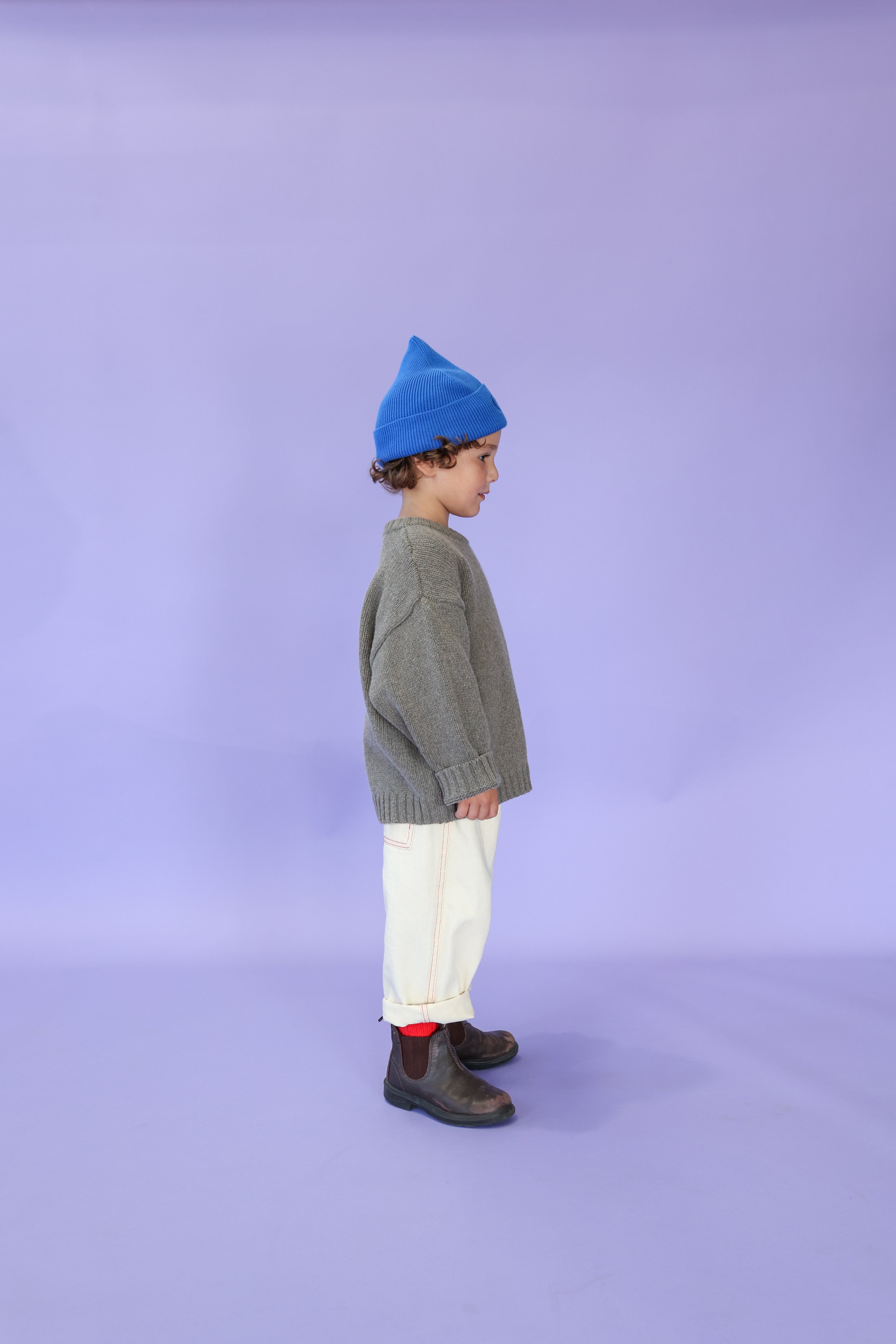 A young child in profile against a purple background wears a blue beanie, gray relaxed fit sweater, GROWN Organic Everyday Denim Pant Milk, red socks, and black boots.
