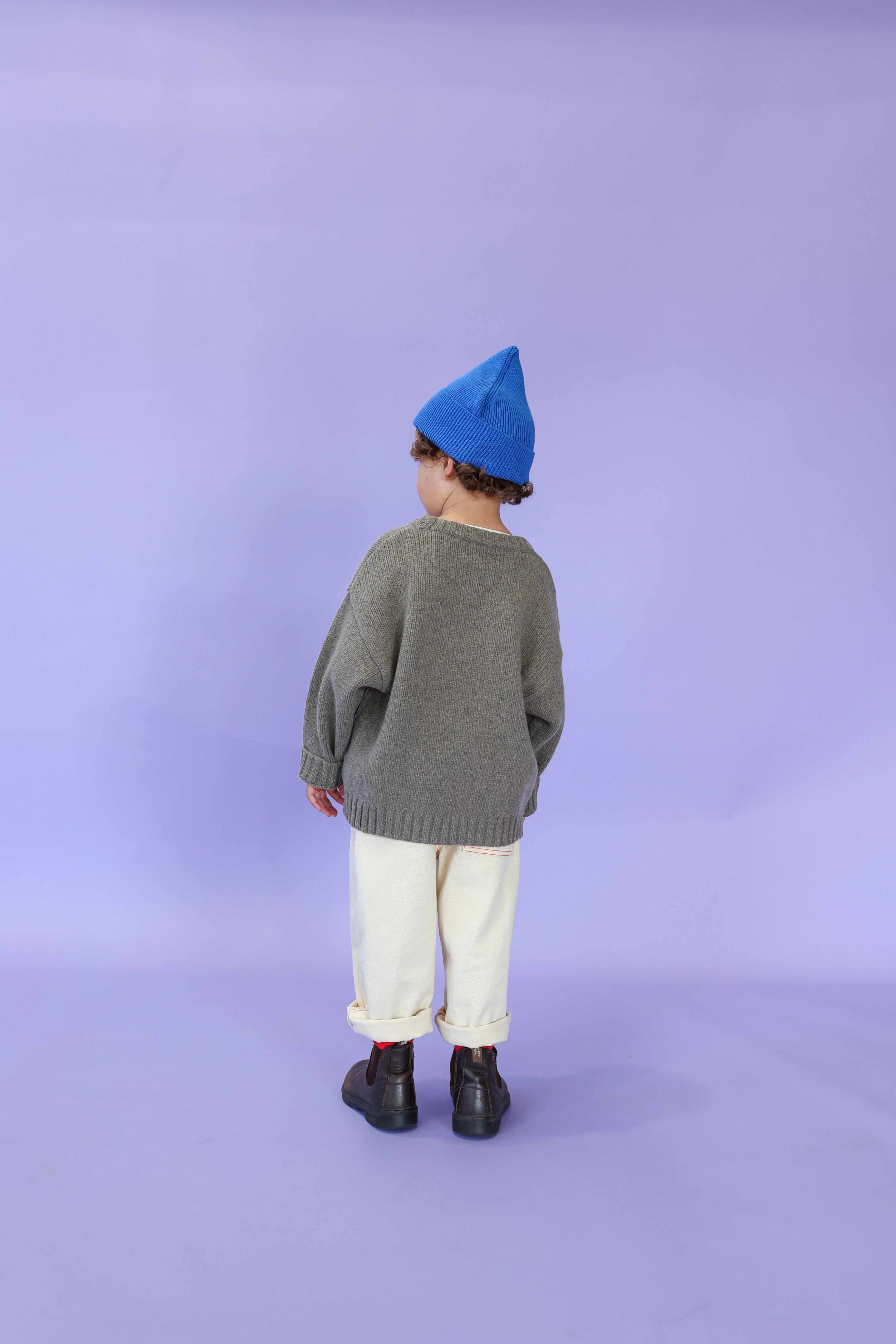 A child stands facing away against a light purple background, wearing a blue beanie, gray organic cotton sweater, black boots, and GROWN's Organic Everyday Denim Pant in Milk.