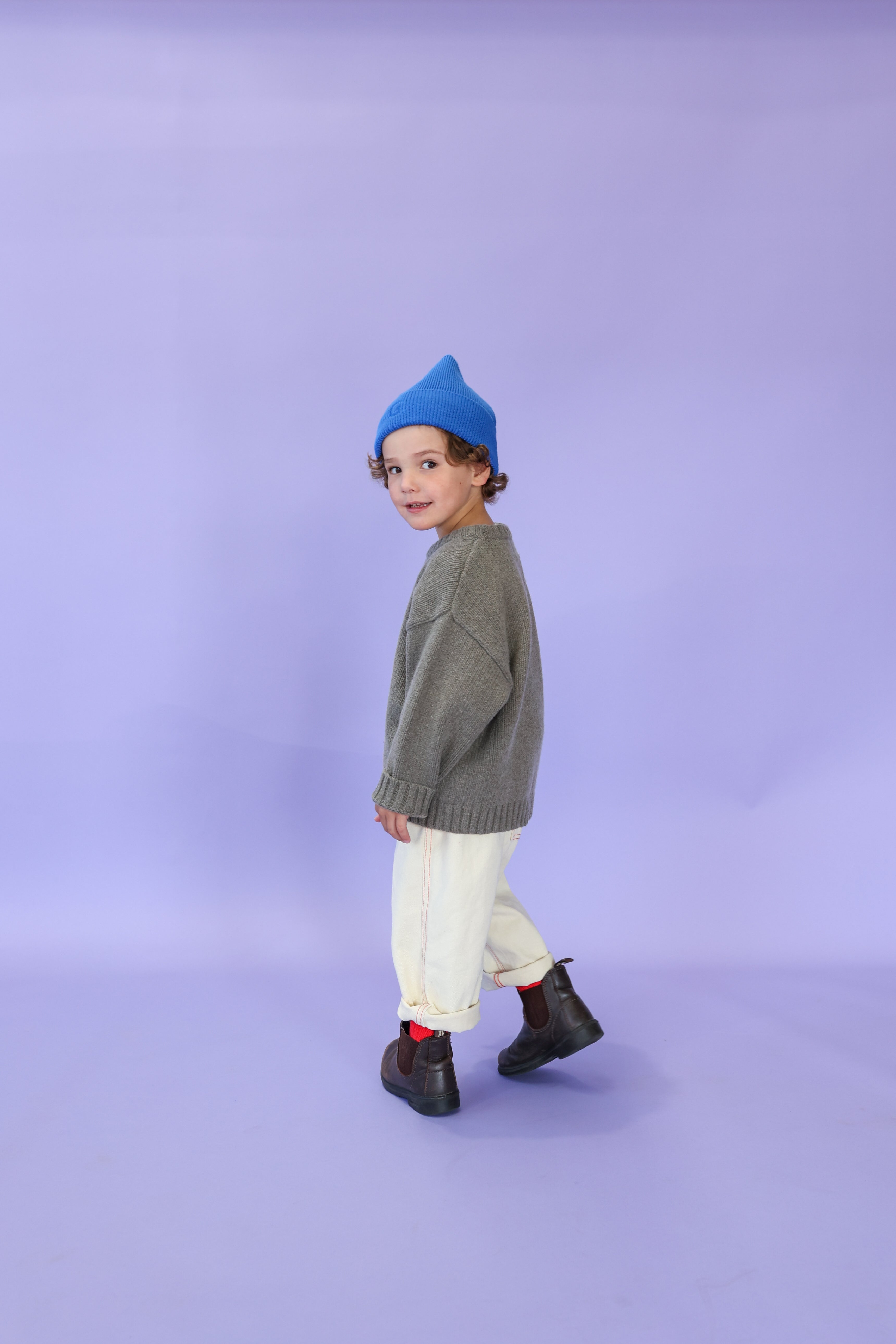 A young child in a blue beanie, gray relaxed fit sweater, GROWN Organic Everyday Denim Pant Milk, and black boots stands against a light purple background, looking back over their shoulder and smiling.