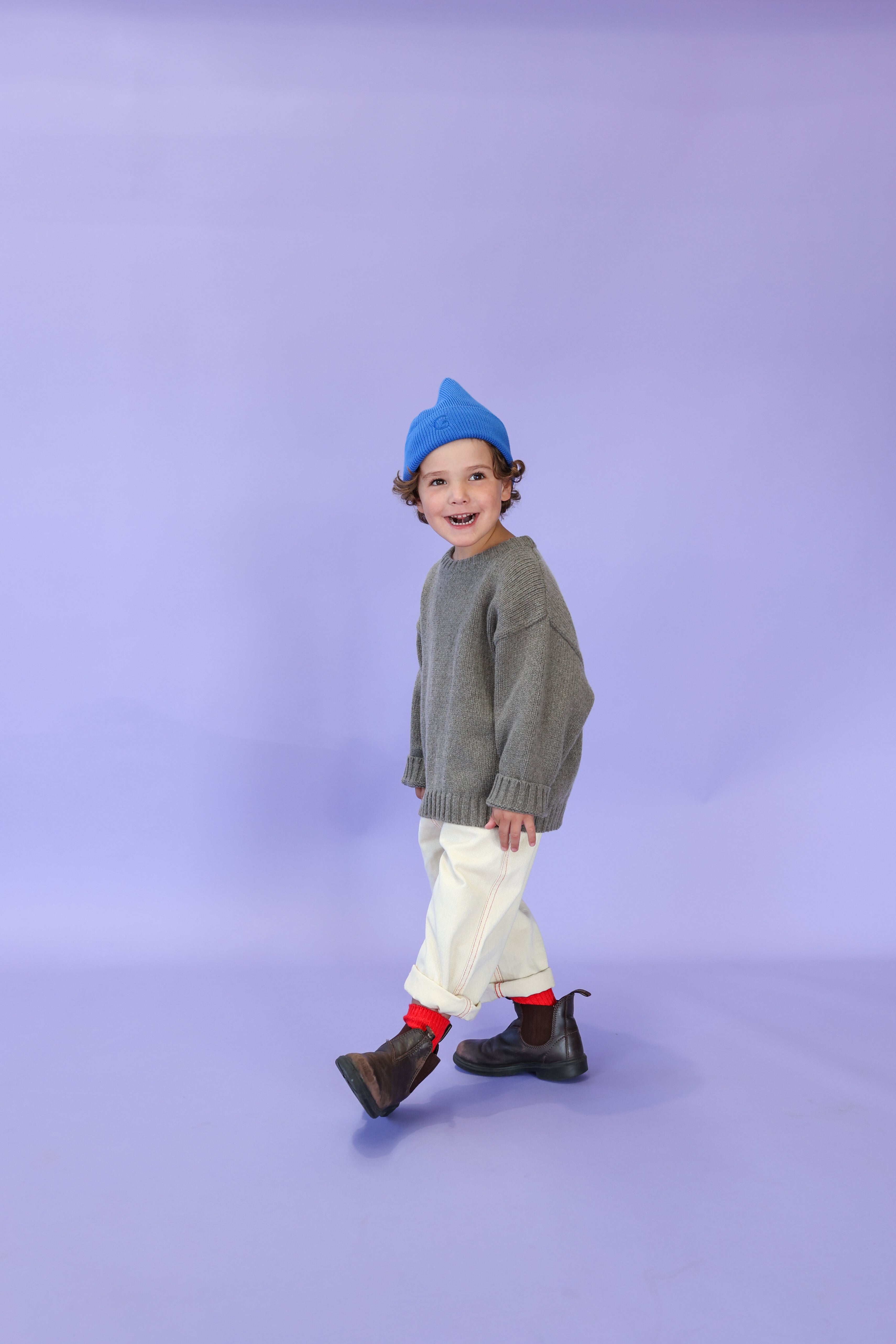 A smiling young child wearing a blue beanie, gray organic cotton sweater, GROWN Organic Everyday Denim Pant Milk, red socks, and brown boots stands against a light purple background.