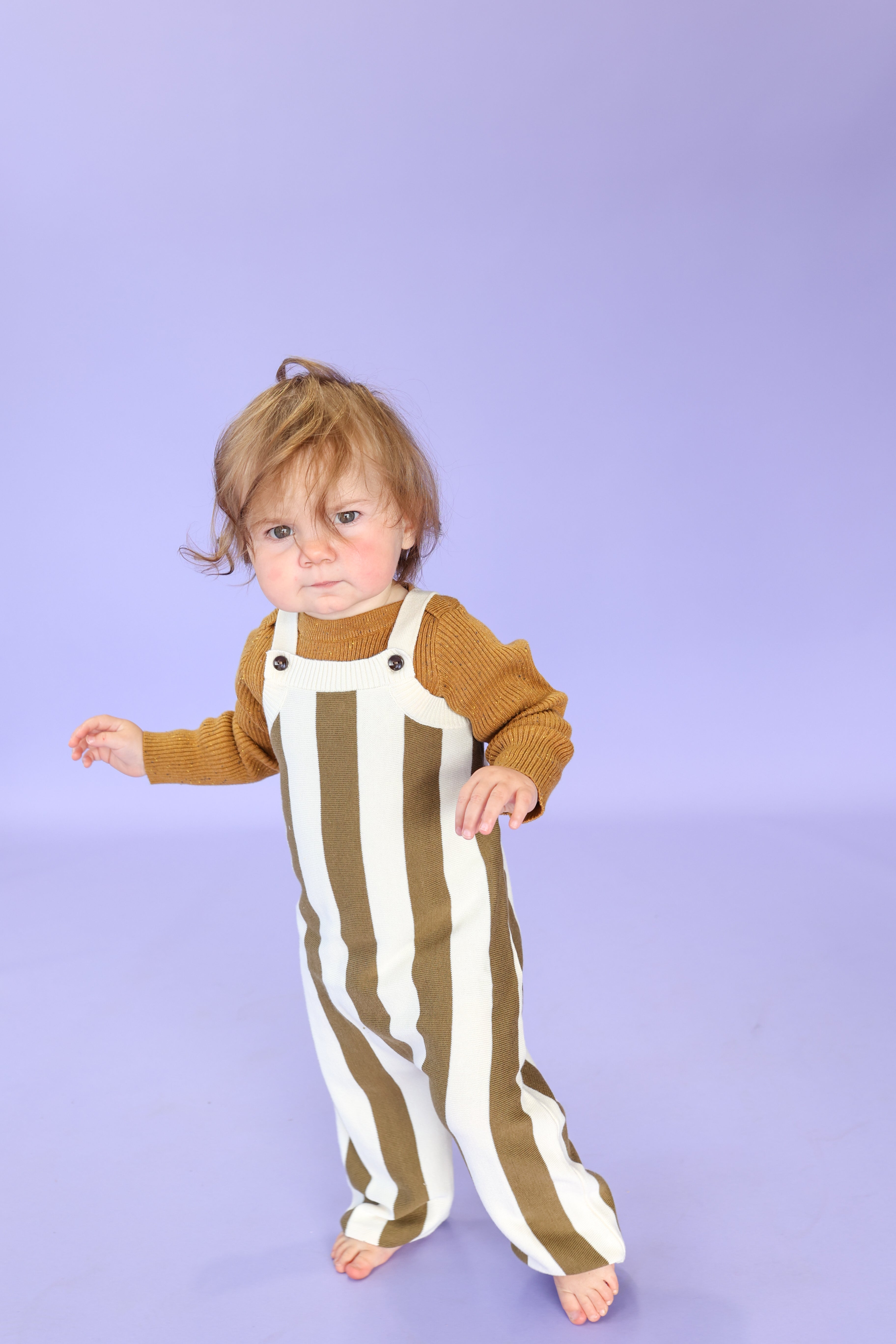 A toddler stands barefoot on a light purple background, wearing GROWN’s Vertical Stripe Knit Overalls—white with bold brown stripes, made from GOTS certified organic cotton, featuring adjustable straps—and a mustard sweater.