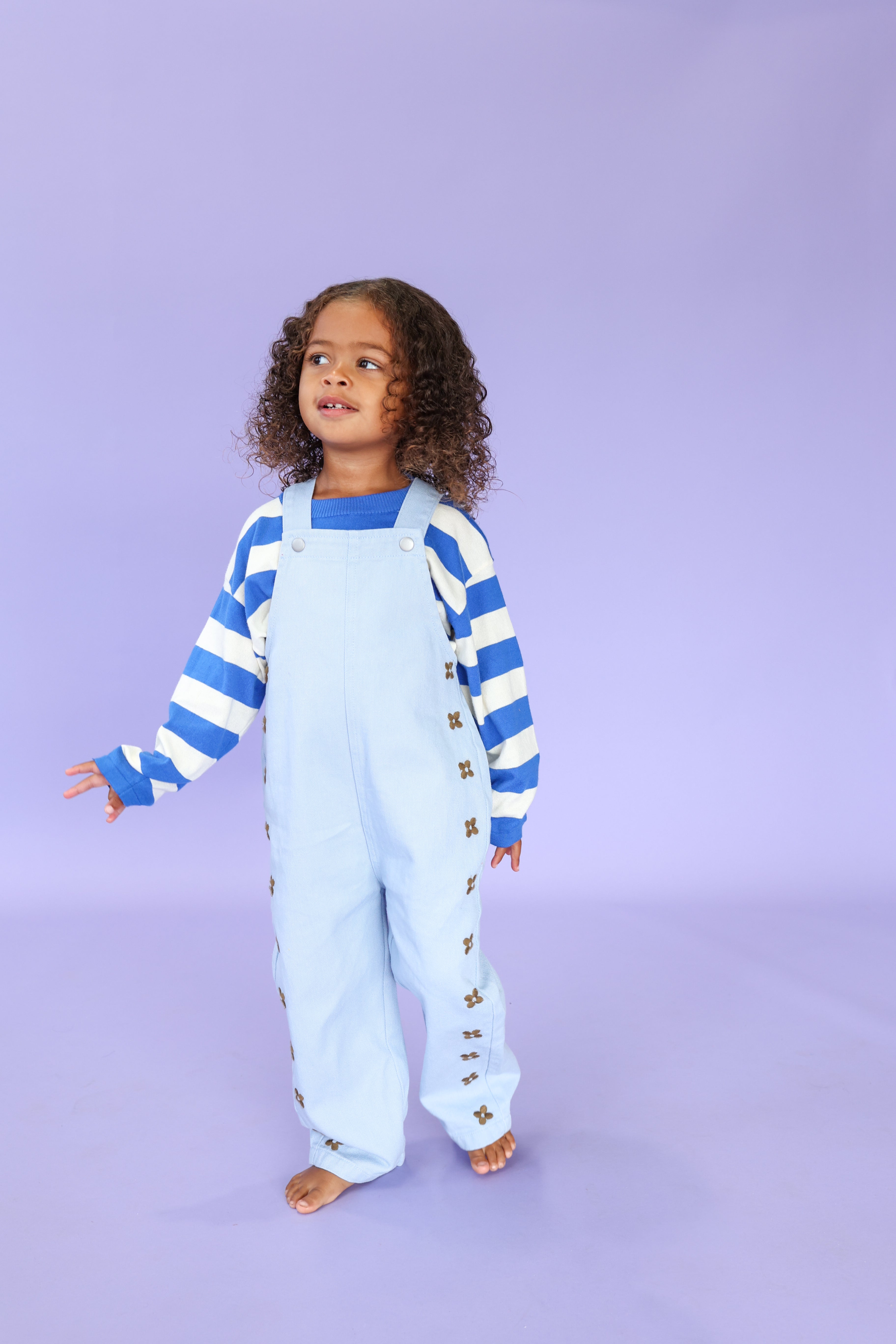 A young child with curly hair stands barefoot on a lavender background, wearing GROWN's Organic Embroidery Posy Overalls over a blue and white striped long-sleeve shirt, looking slightly to the side.