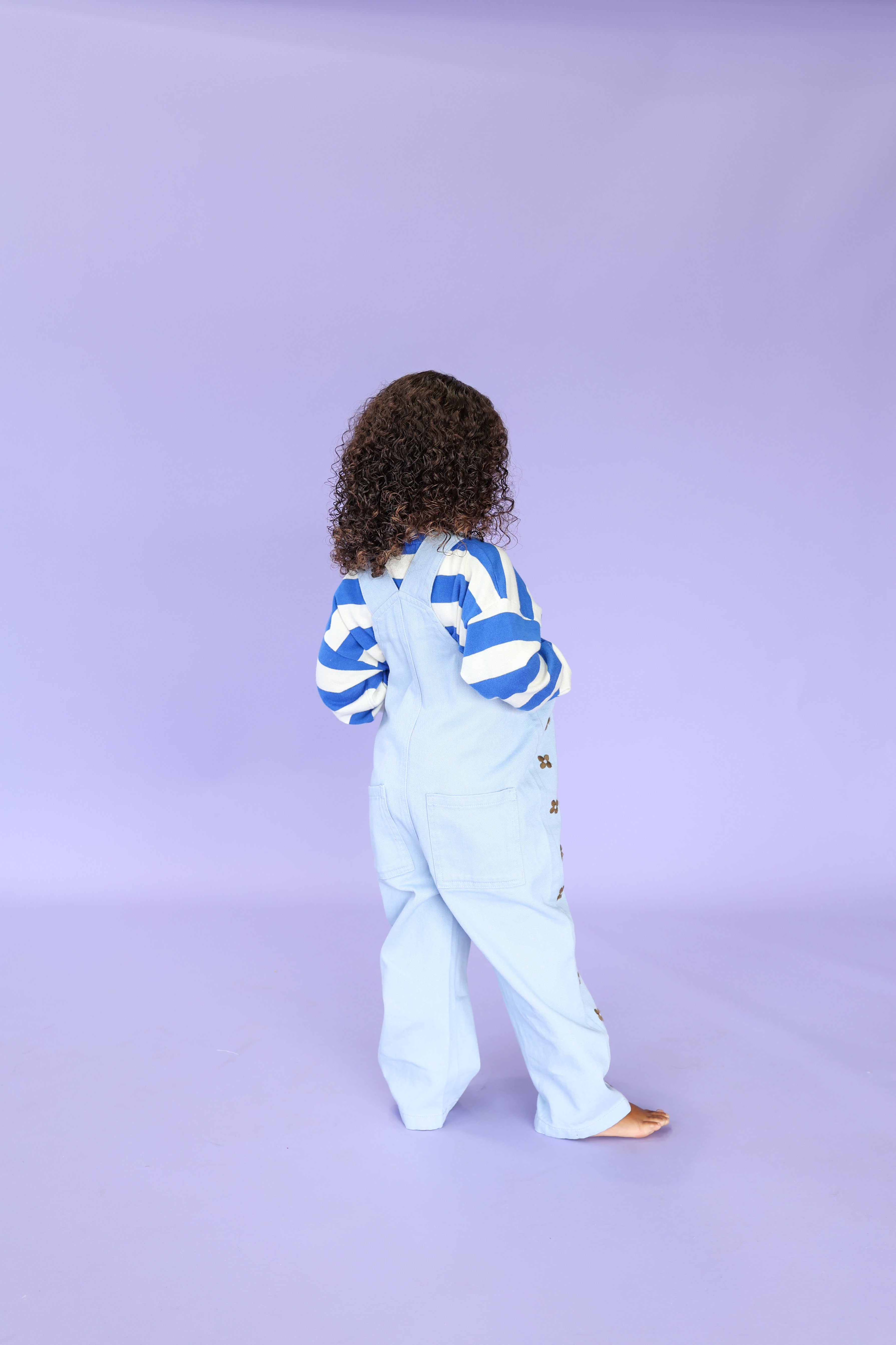 A young child with curly hair, barefoot, wears GROWN's Organic Embroidery Posy Overalls in light blue denim, featuring posy embroidery and metal press snaps, and stands facing away against a solid light purple background.