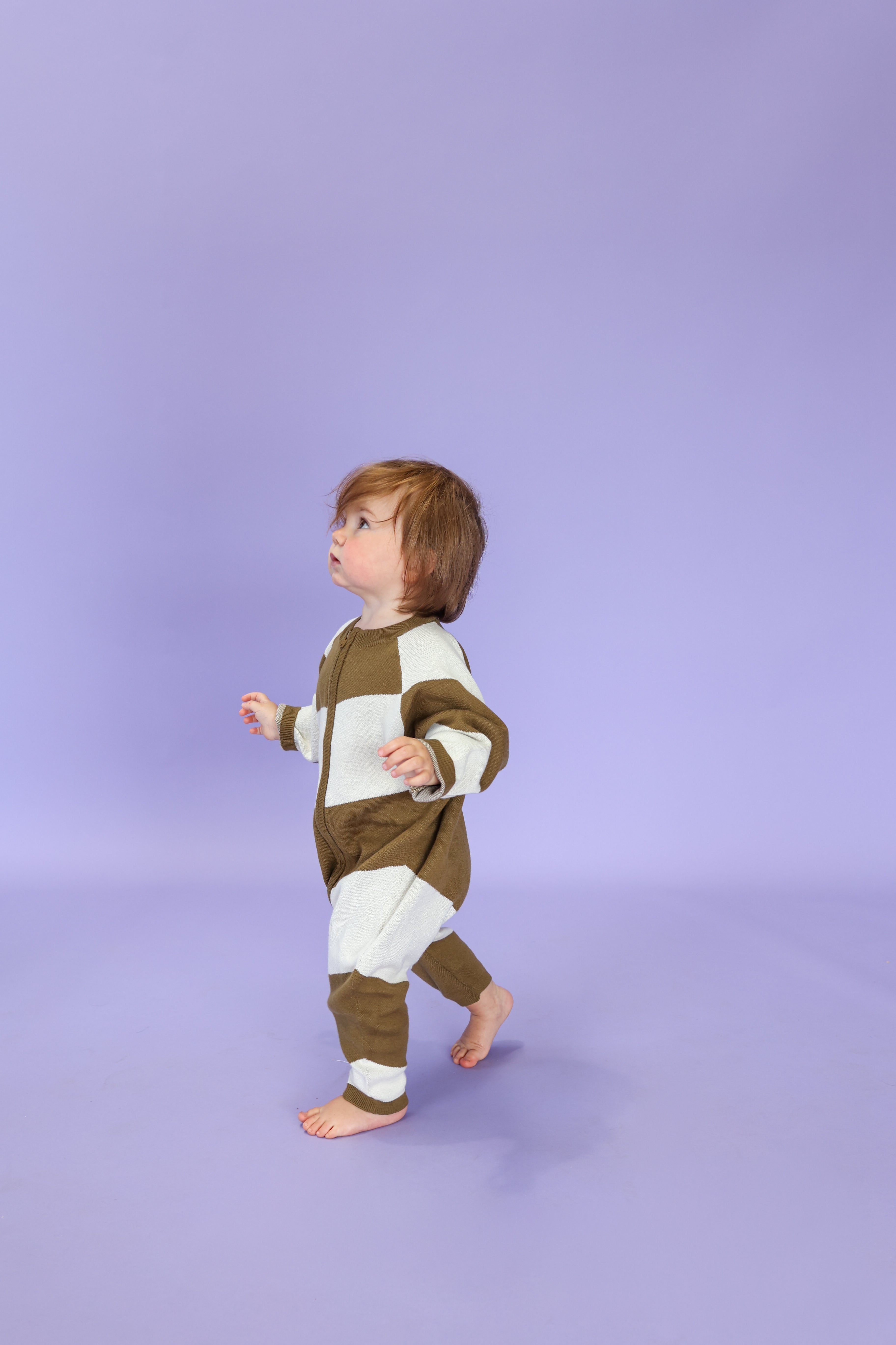 A toddler with brown hair wears the GROWN Organic Essential Zip Starsuit in Coffee Stripe and walks barefoot on a lavender floor with a matching background, looking upwards.