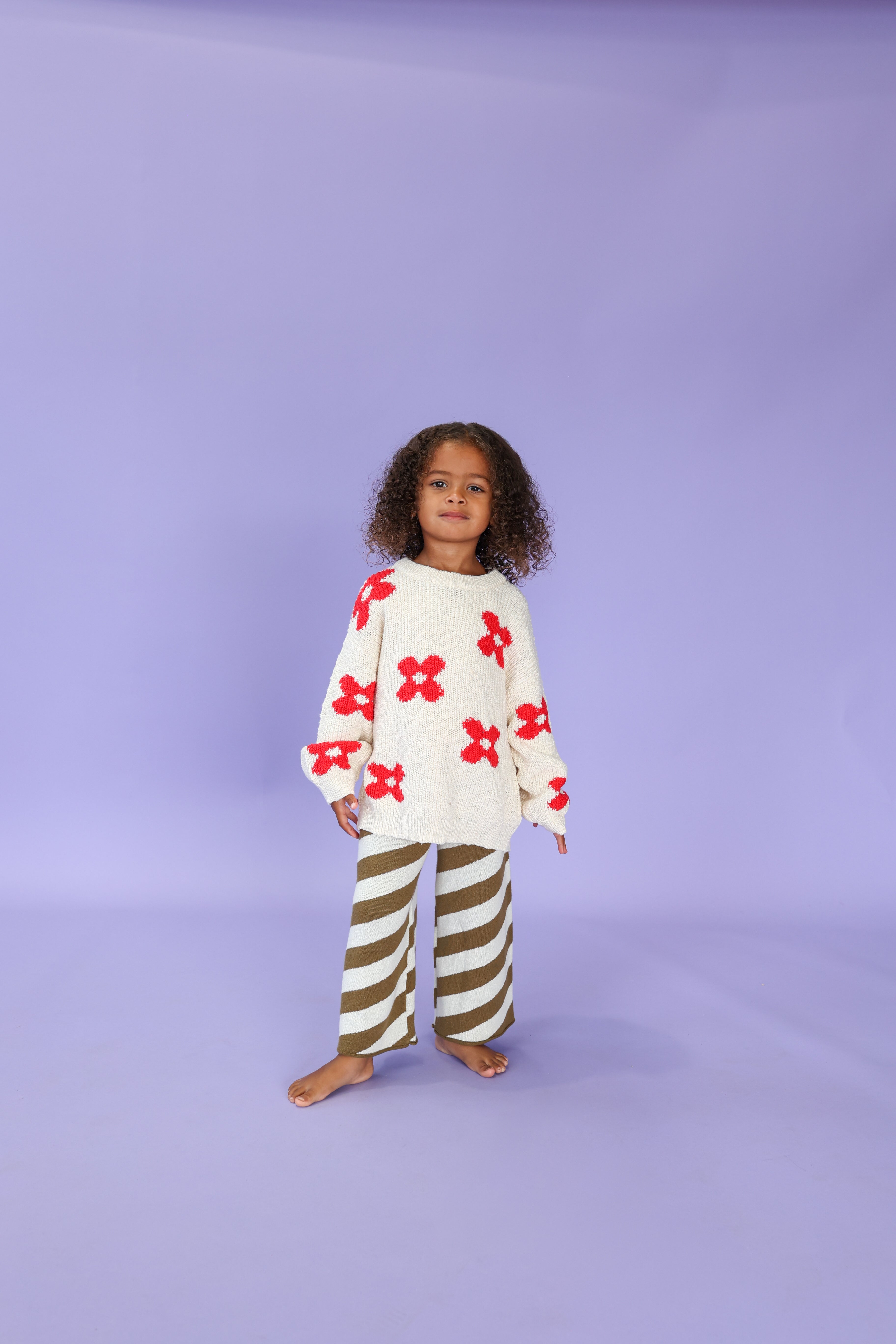A young child with curly hair stands barefoot on a light purple background, wearing a white sweater with red flower patterns and GROWN Diagonal Stripe Knit Flares featuring an elastic waistband.