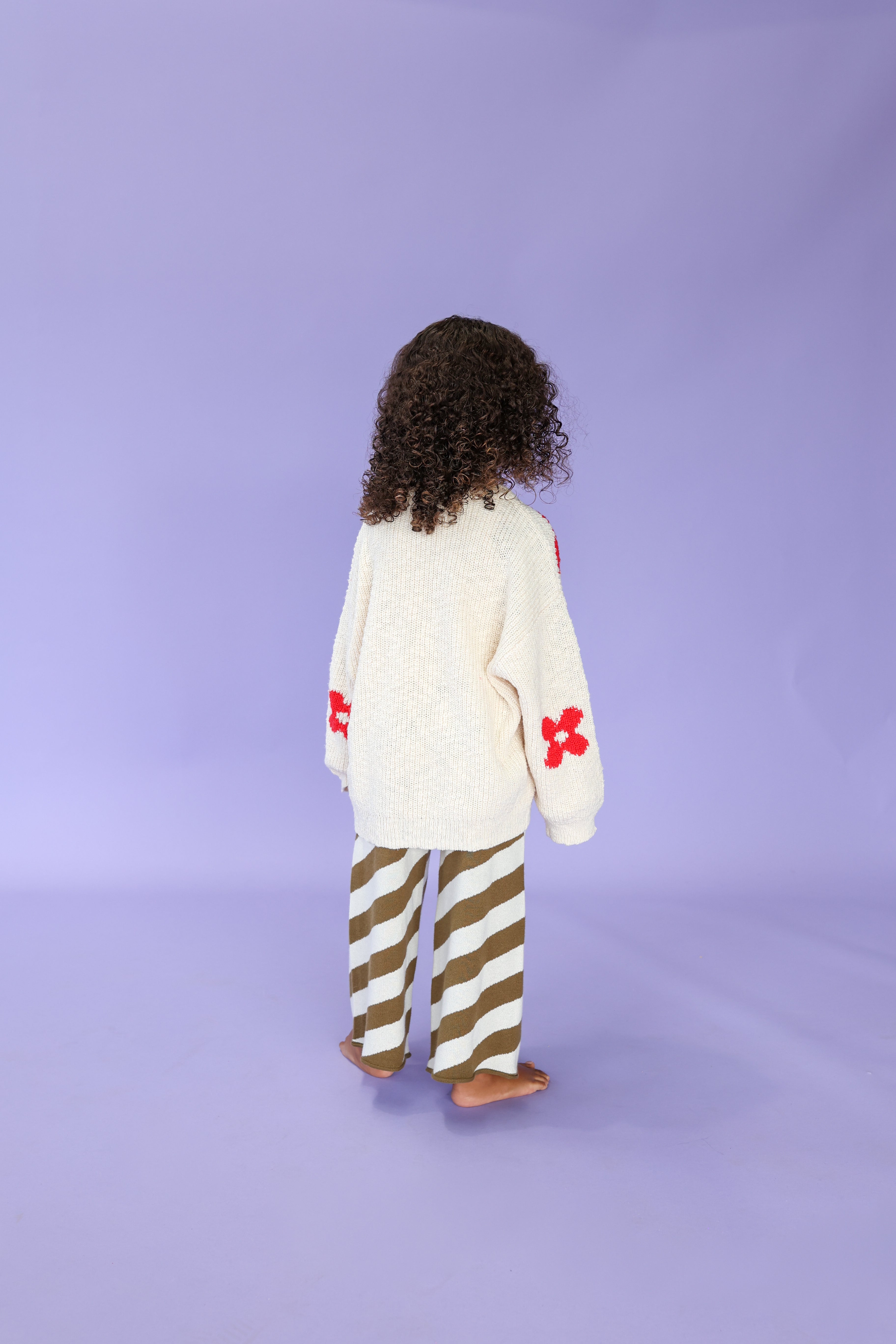 A child with curly hair stands barefoot on a purple floor, facing away. They wear a cream-colored sweater with red flower designs on the sleeves and GROWN - Diagonal Stripe Knit Flares by GROWN.