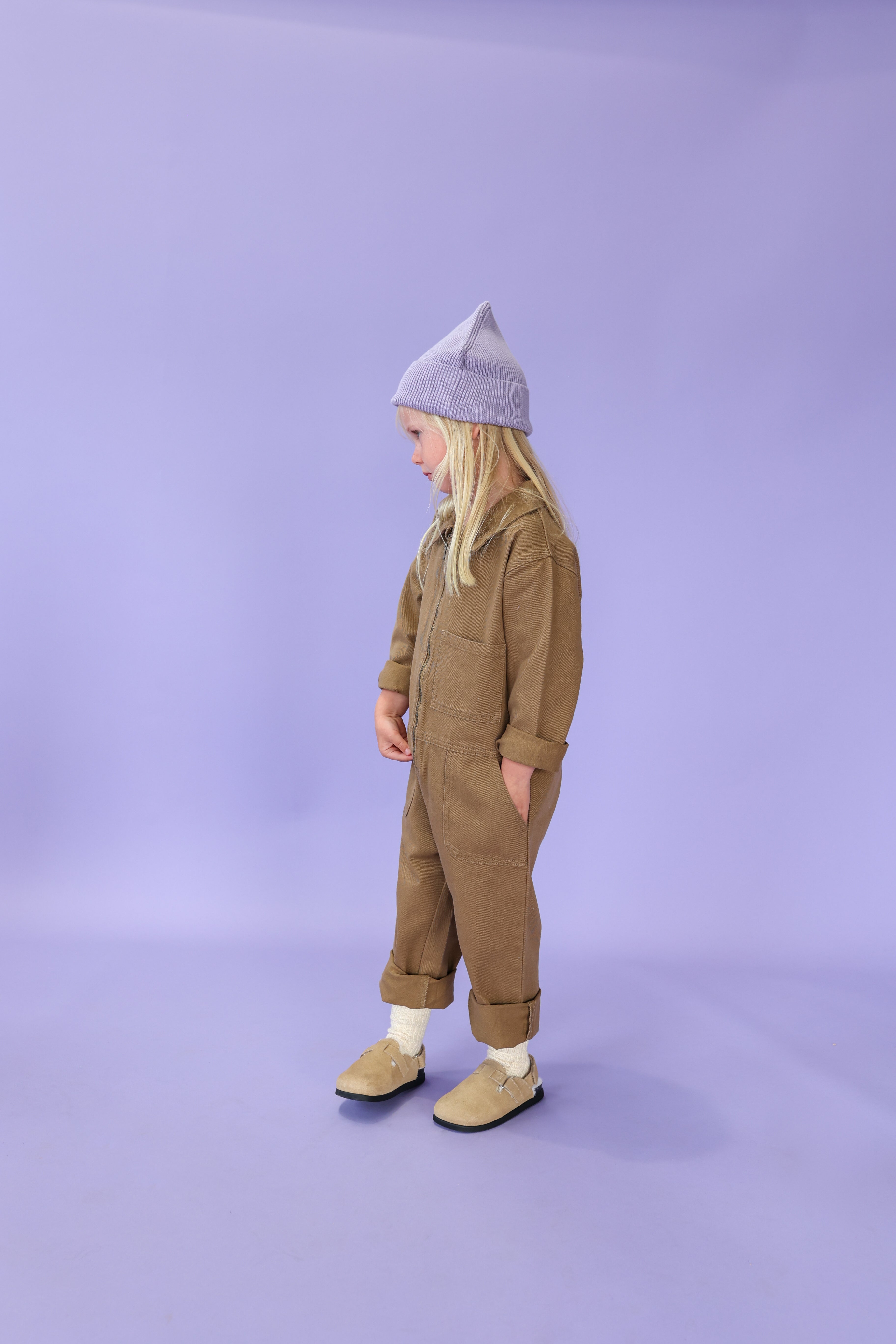 A young child with long blonde hair stands against a light purple background, looking downward while wearing a tan jumpsuit, cream socks, tan slip-on shoes, and the GROWN Organic Ribbed Super Pixie Beanie in Mauve.