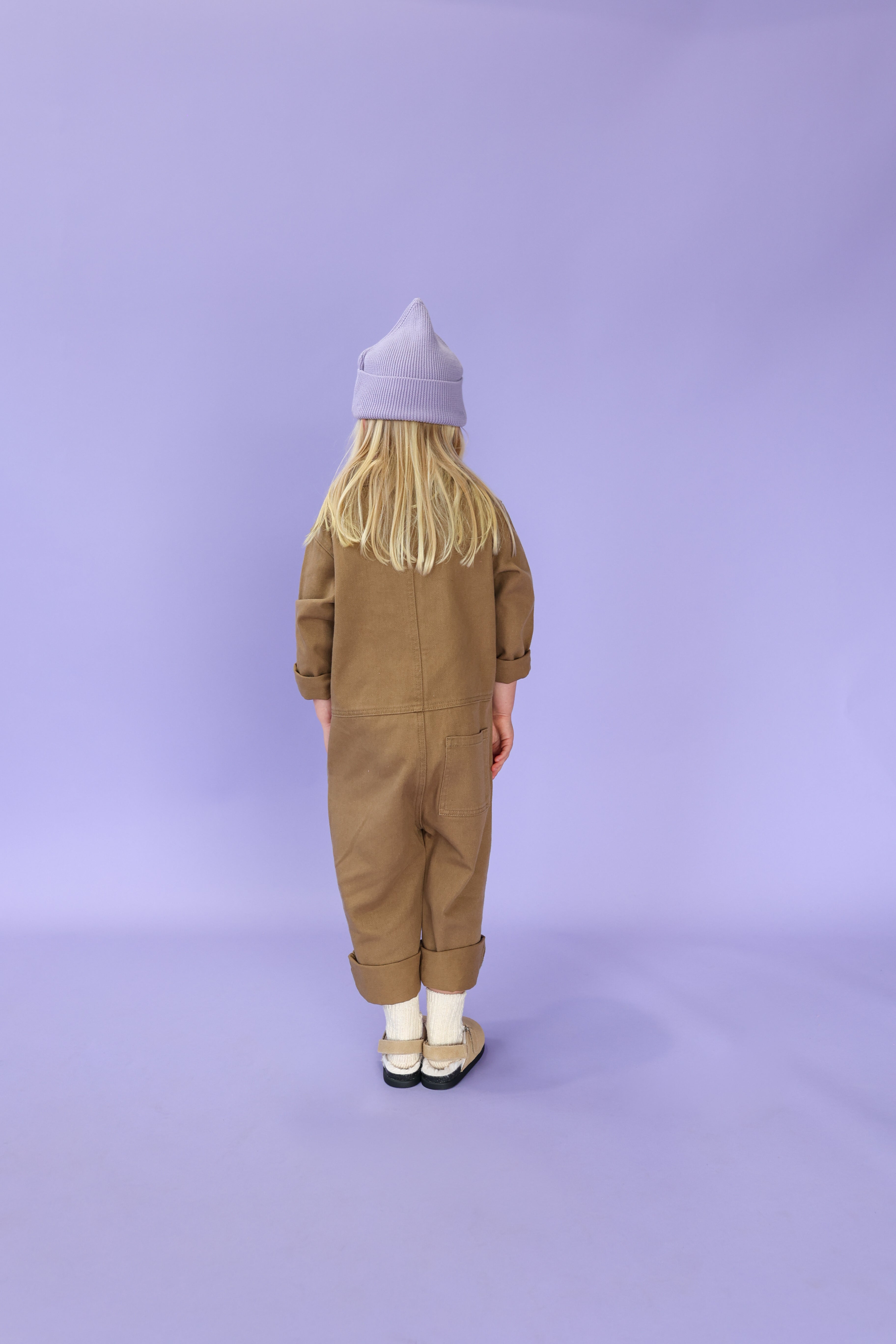 A young child with long blond hair, wearing the GROWN Organic Ribbed Super Pixie Beanie in Mauve, a brown jumpsuit, cream socks, and white organic cotton shoes, stands facing away against a plain lavender background.
