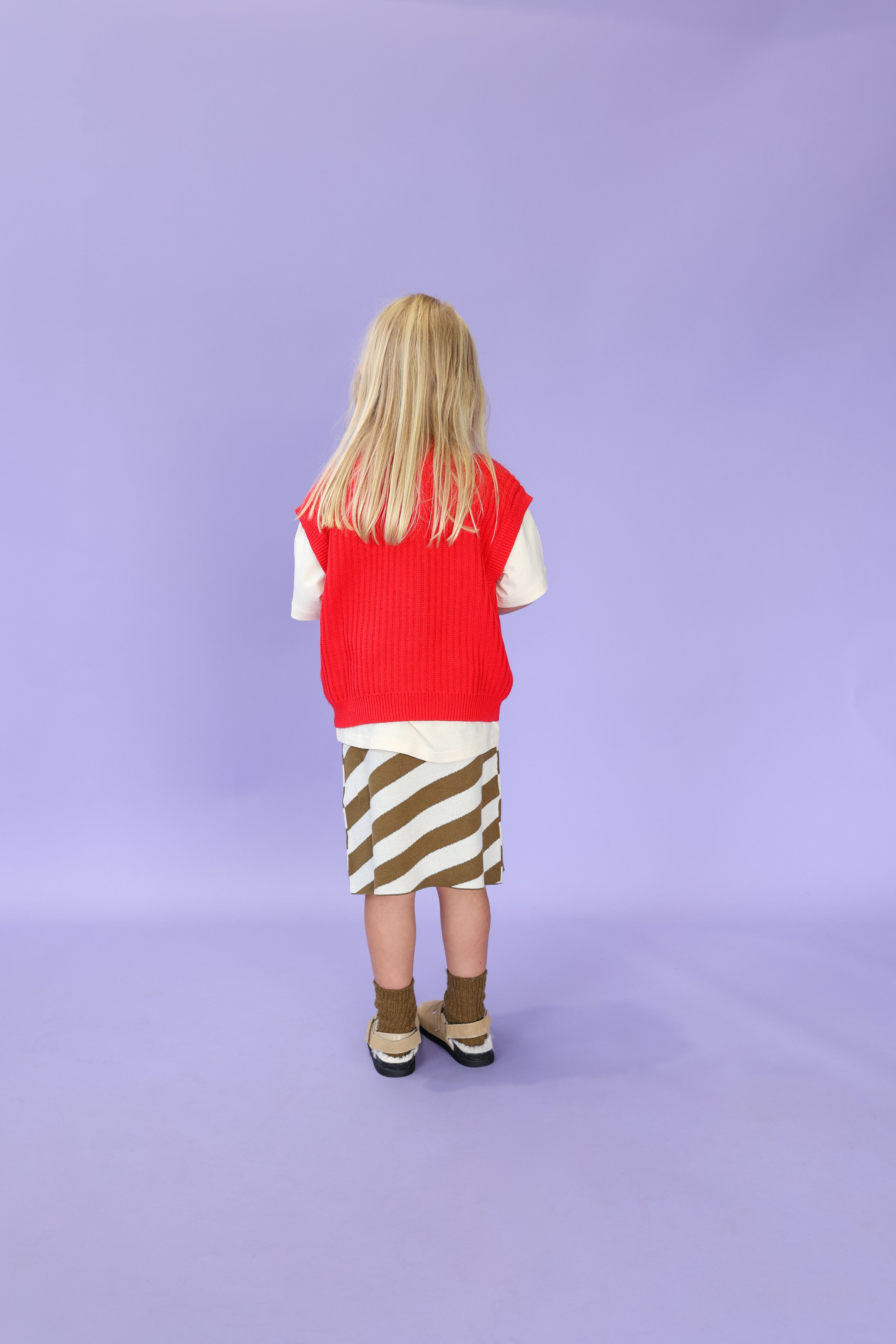 A young child with long blonde hair, facing away, wears the GROWN Chunky Rib V-Neck Vest in Flame over a white organic cotton shirt, a brown and white striped skirt, brown socks, and sandals against a light purple background.