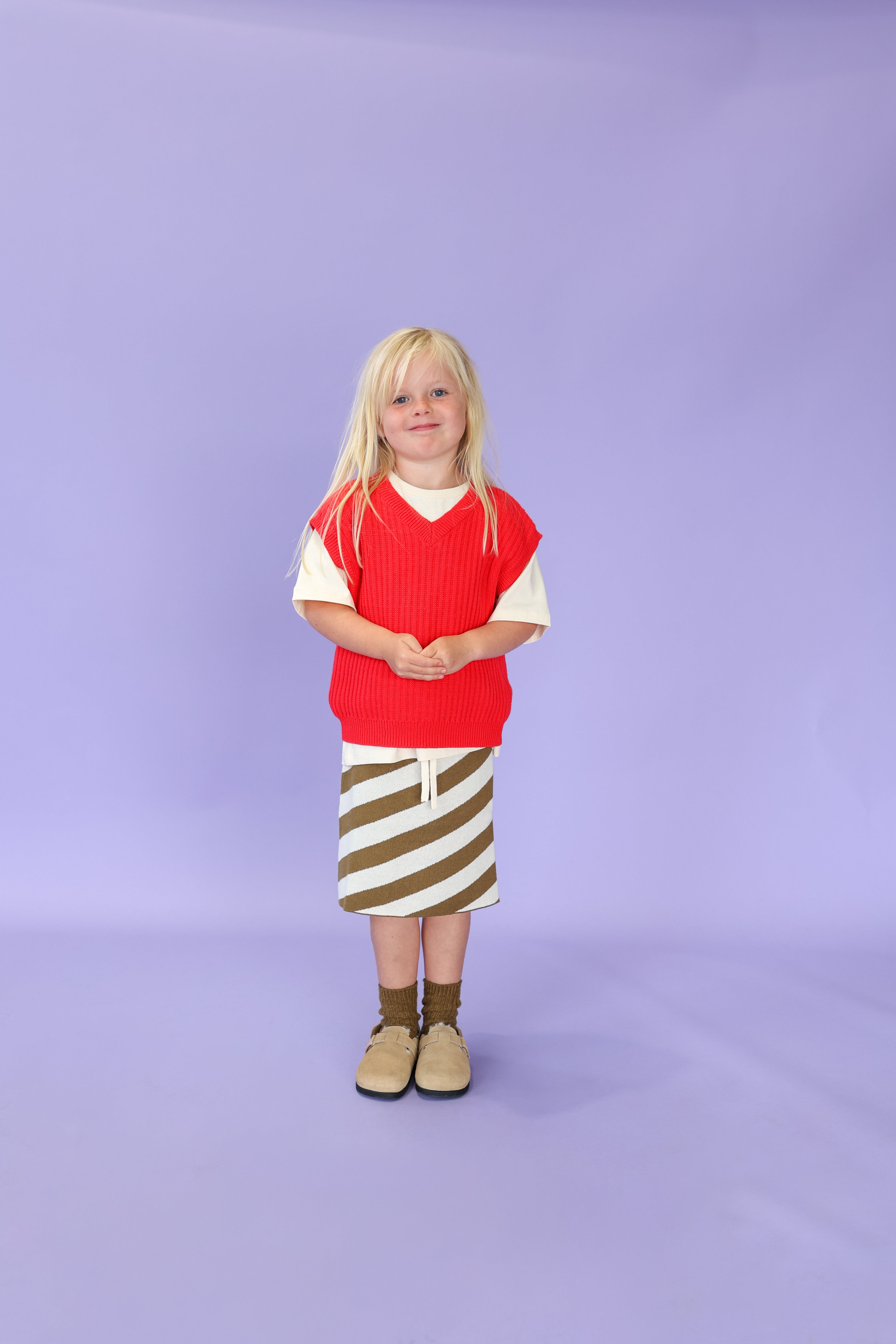 A young child with long blonde hair stands against a light purple backdrop, wearing the GROWN Chunky Rib V-Neck Vest Flame over a white shirt, paired with a brown and white striped skirt, brown socks, and beige shoes.