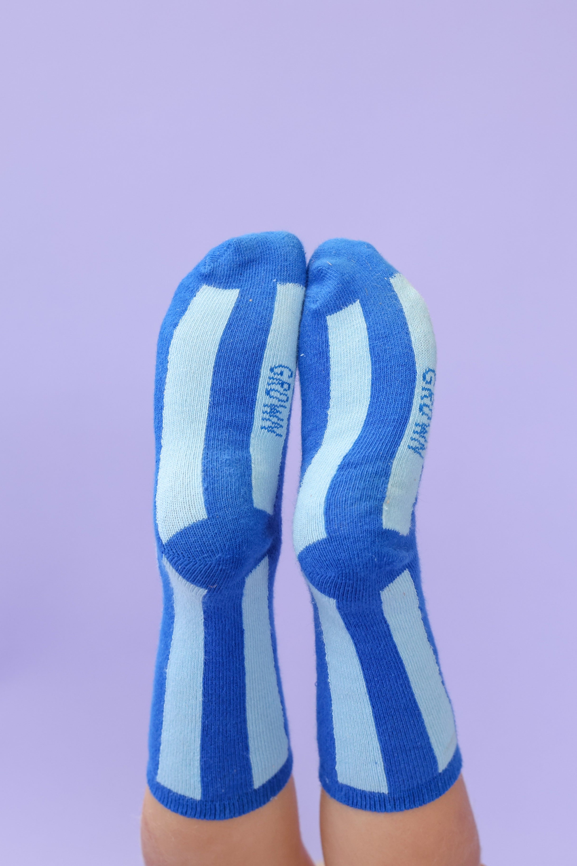 Two feet wearing GROWN - Striped Socks Sea are held together, pointing upward against a plain light purple background.