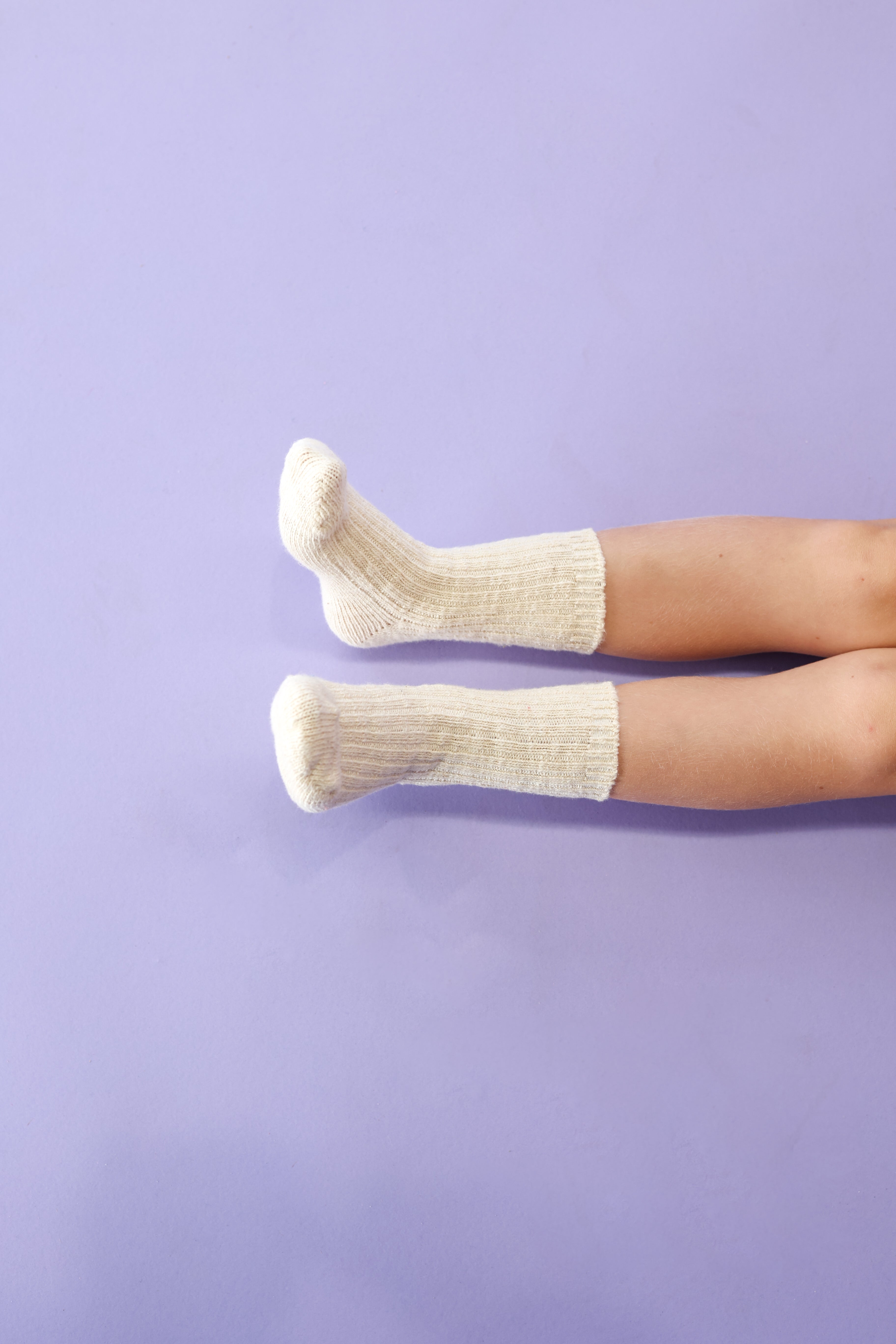 A pair of legs wearing GROWN's Grown - Chunky Rib Sock Milk is resting on a light purple surface, with feet slightly elevated and toes pointed upward.