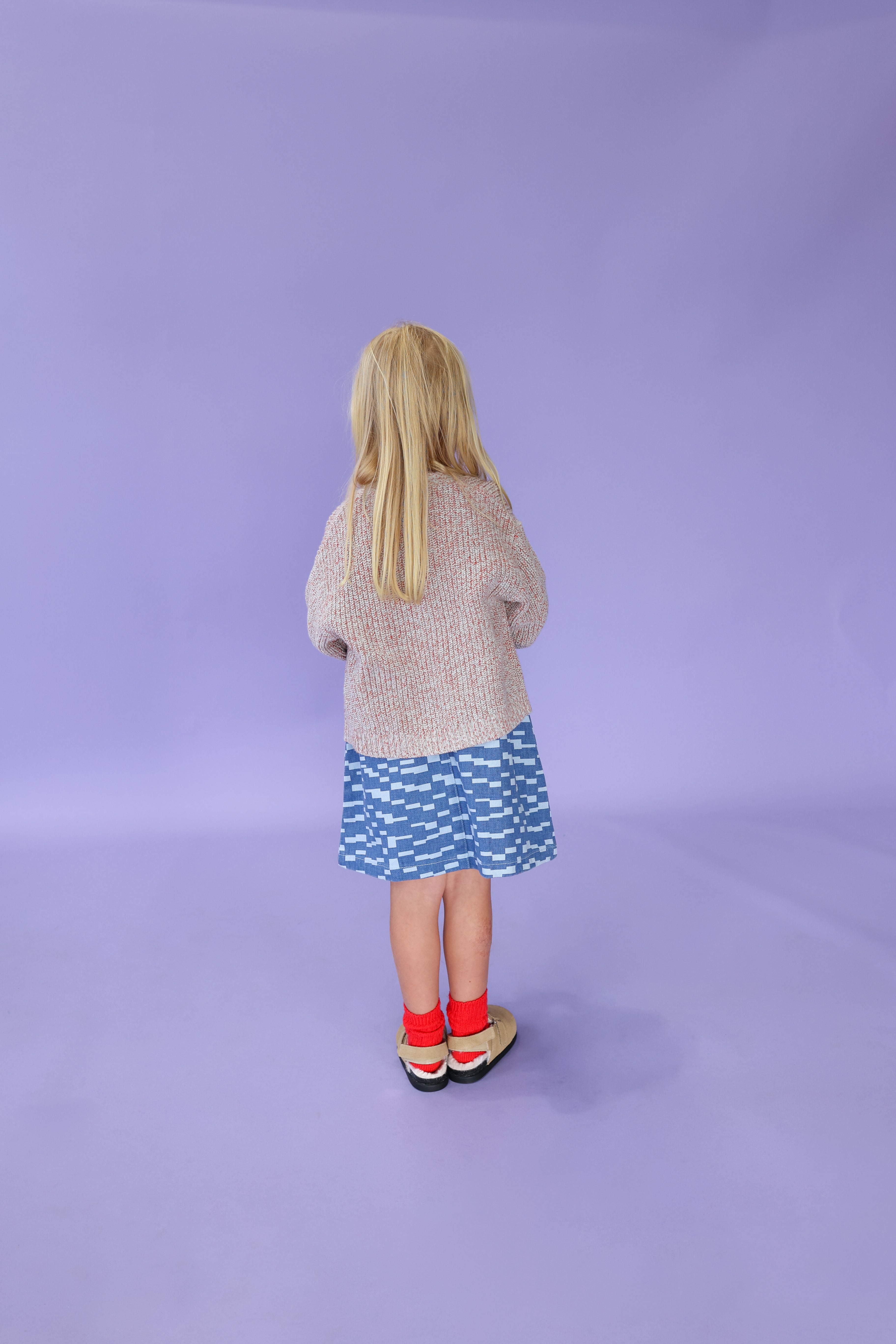 A young girl with long blonde hair wears the GROWN Twisted Chunky Rib Cardigan, a relaxed fit sweater, along with a blue patterned skirt, bright red socks, and mismatched shoes, standing against a plain light purple background.