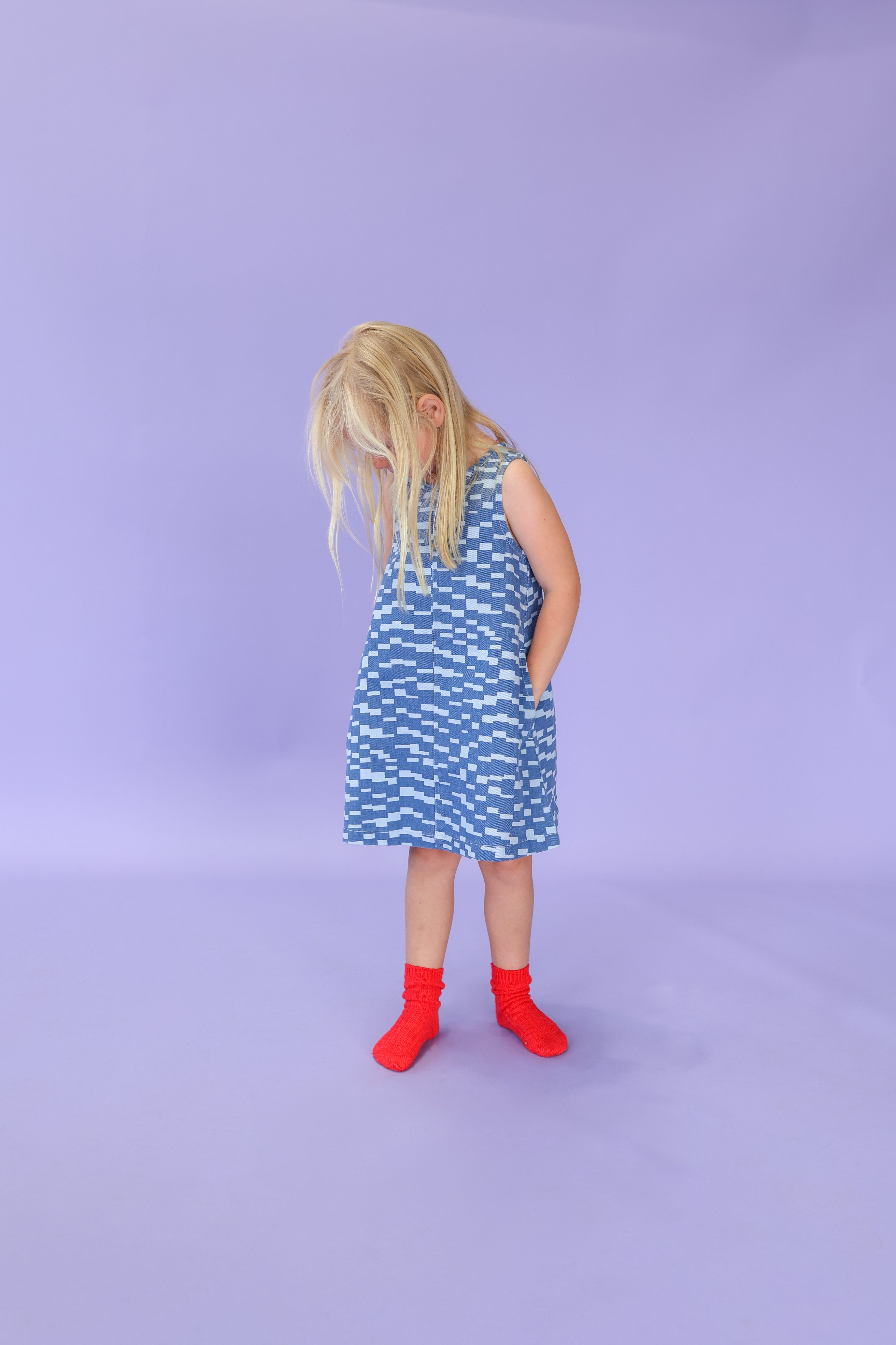 A young girl with blonde hair stands against a light purple background, looking down with her hands in her pockets, wearing the GROWN Boxes Denim Dress and bright red socks.