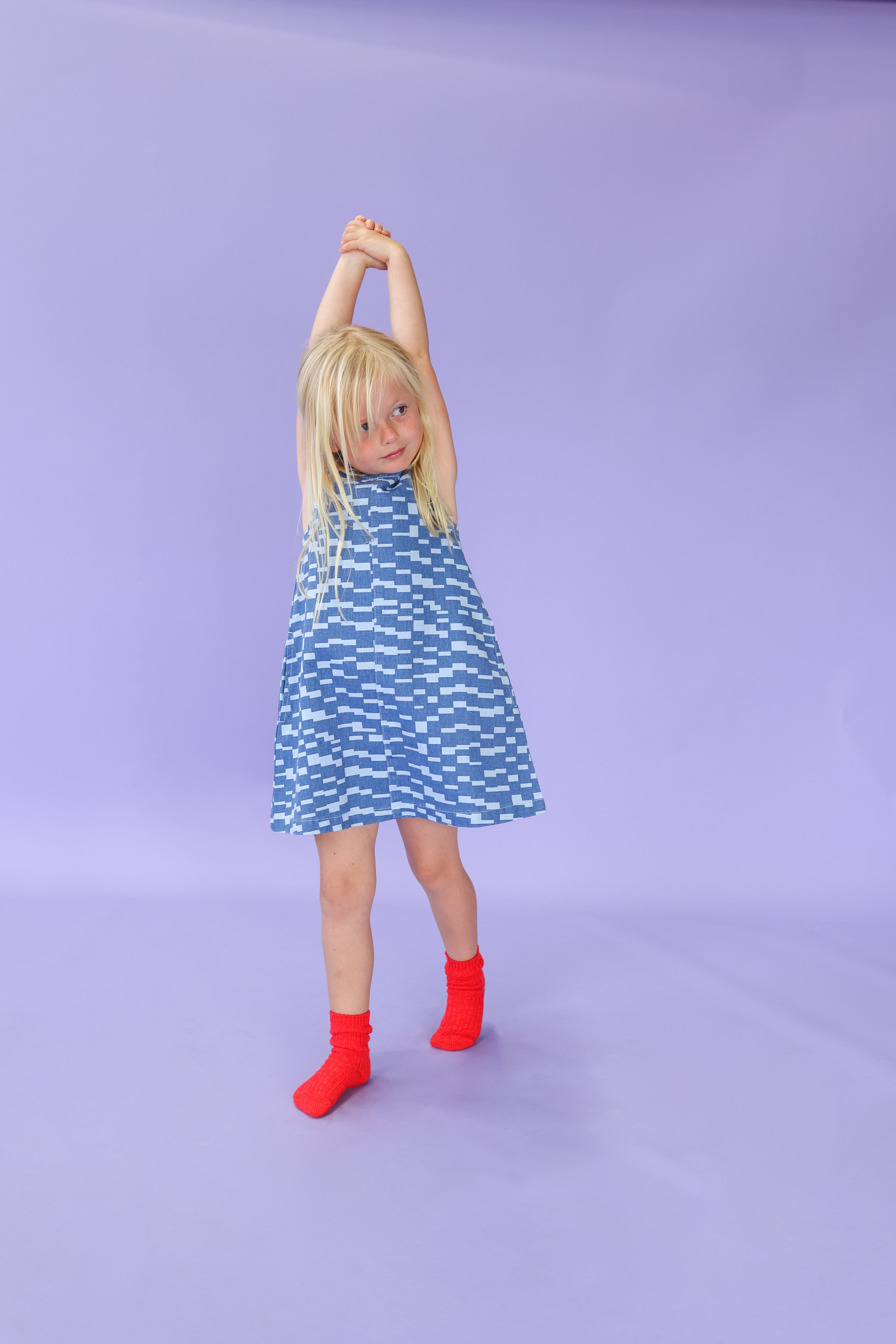 A young girl with blonde hair wears the GROWN Boxes Denim Dress, stretching her arms above her head against a light purple background.