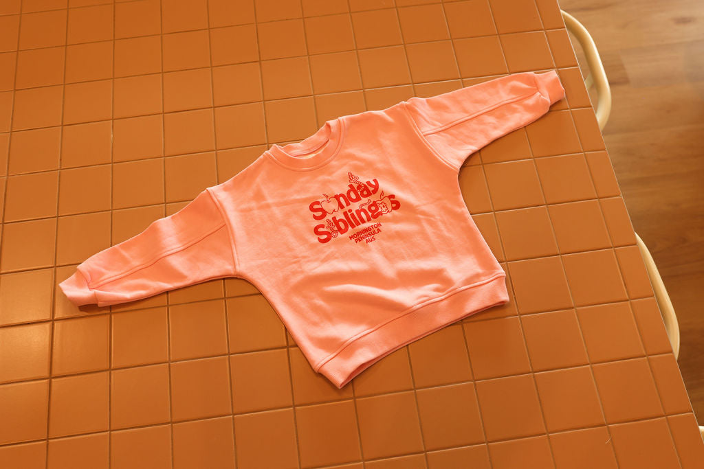 A light pink Doc Sweater Pink from SUNDAY SIBLINGS, featuring the words "Sunday Siblings" in red text, is neatly laid out on a tiled surface. Made from high-quality French terry cotton, its relaxed oversized fit ensures casual comfort, making it perfect for any laid-back day.