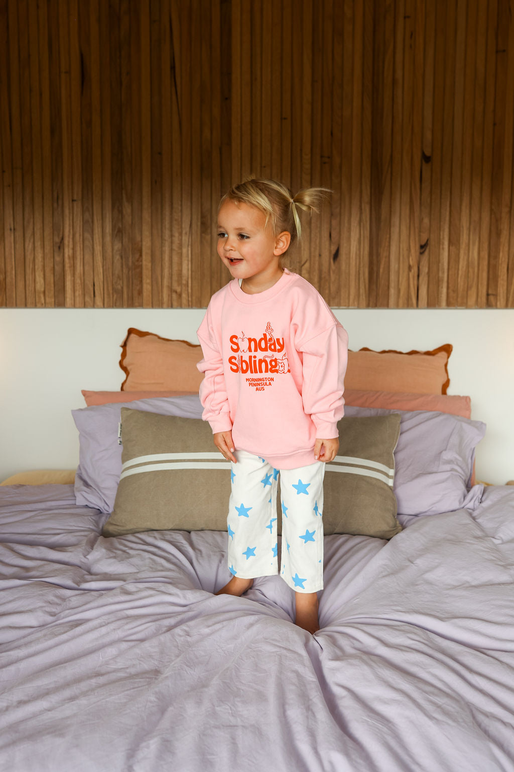 A cheerful young child with blond hair, wearing the Doc Sweater Pink made of high-quality French terry cotton from Sunday Siblings and star-patterned pajamas, stands on a bed with a wooden headboard and light purple bedding.